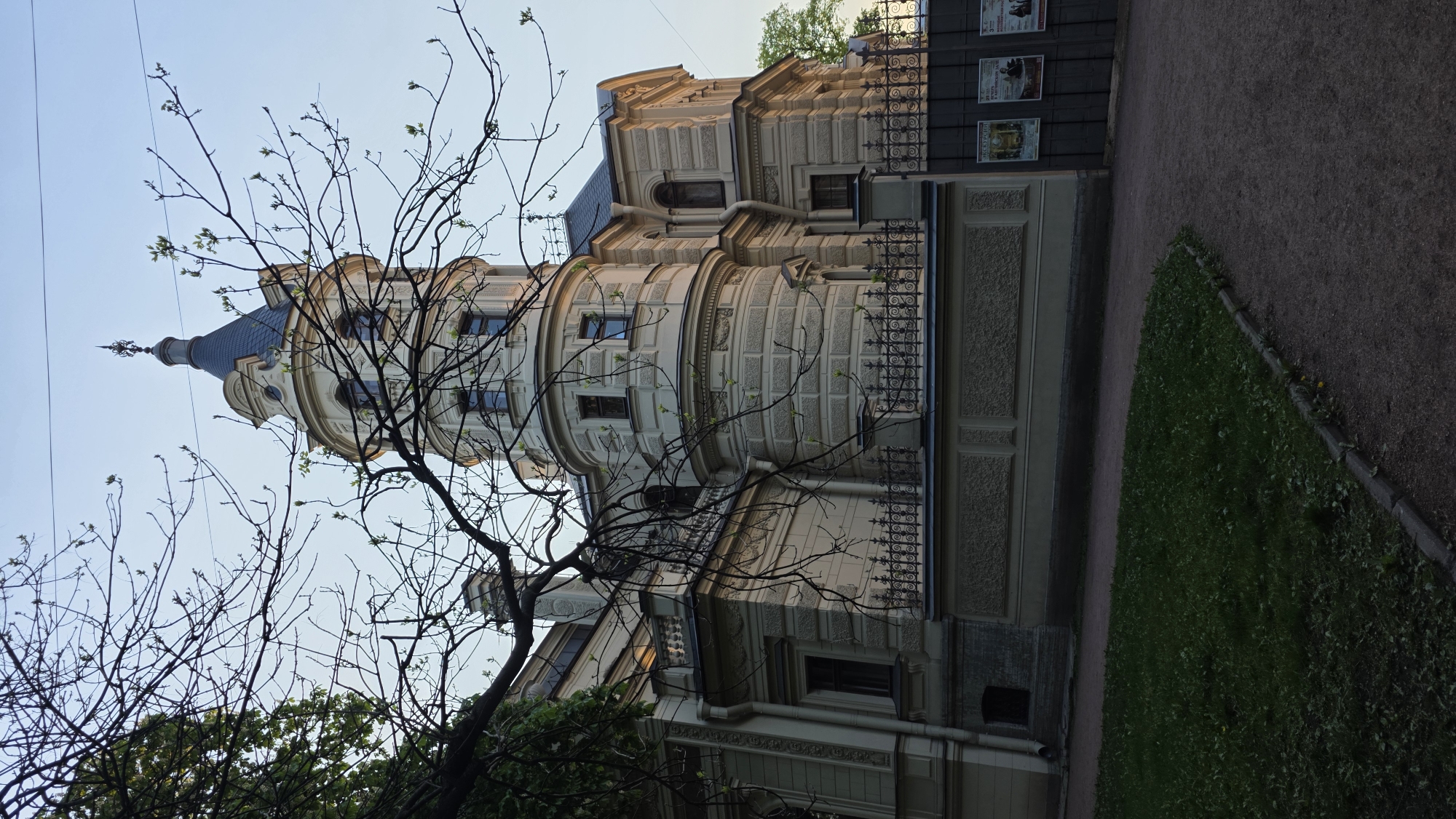 The image depicts a building with a prominent, round tower and a blue, pointed roof in Saint Petersburg, Russia. The building's exterior is ornate, featuring detailed masonry work, decorative windows, and a wrought-iron fence. A bare tree with budding leaves stands in the foreground, partially obscuring the building. A narrow path runs along the side of the building, separating it from a grassy area. Posters or signs are visible on a wall behind the fence. The sky is light, suggesting either early morning or late afternoon.