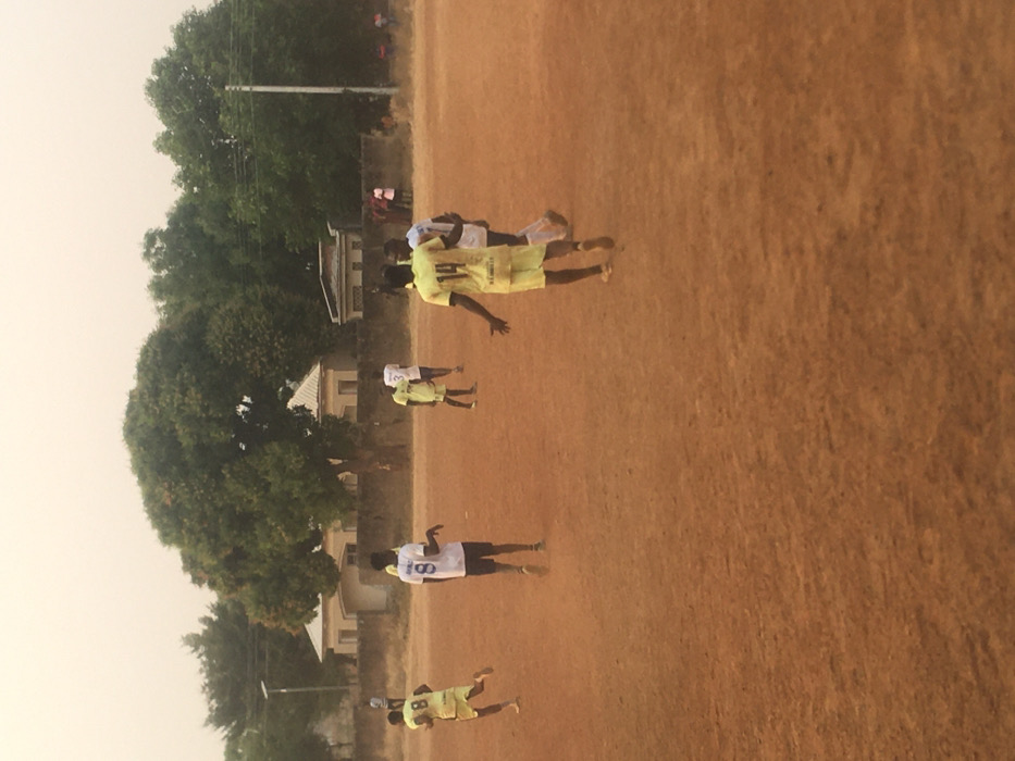 A low-angle shot shows several people on a reddish-brown field, likely playing a game. They are wearing light-colored jerseys with numbers. The player closest to the camera wears number 8. Trees line the edge of the field, and some buildings are visible in the background. The sky is hazy. The image is slightly rotated counter-clockwise.
