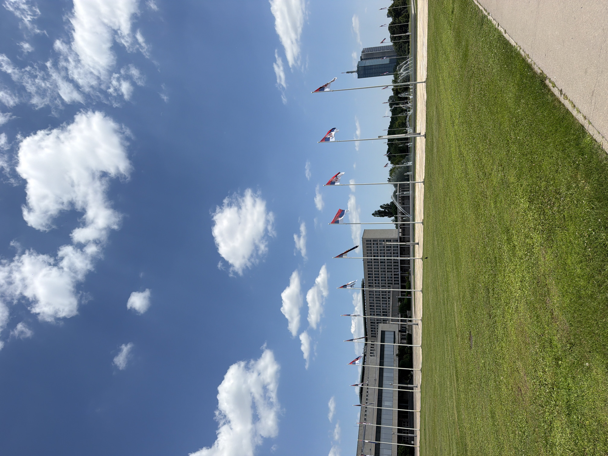 A wide, low-angle shot captures a bright, sunny day in Belgrade, Serbia, with fluffy white clouds scattered across a vibrant blue sky. In the foreground, a neatly trimmed green lawn stretches across the scene, bordered on the right by a concrete path. Behind the lawn, a row of tall flagpoles fly numerous flags, many of which appear to be the Serbian flag with its distinctive red, blue, and white horizontal stripes and crest.
Dominating the midground is a long, imposing building with a symmetrical facade of windows and concrete columns. This building is identified as the Palace of Serbia, a significant administrative complex located in New Belgrade. To the right of the Palace of Serbia, a modern, glass-faced skyscraper stands tall, hinting at the urban landscape of the city. Further in the distance, multiple fountains are visible, adding a touch of elegance to the open plaza. The overall scene suggests a formal and monumental public space, possibly the Sava Centar or a similar area in New Belgrade known for its grand architecture and open spaces. No people are visible in the frame.