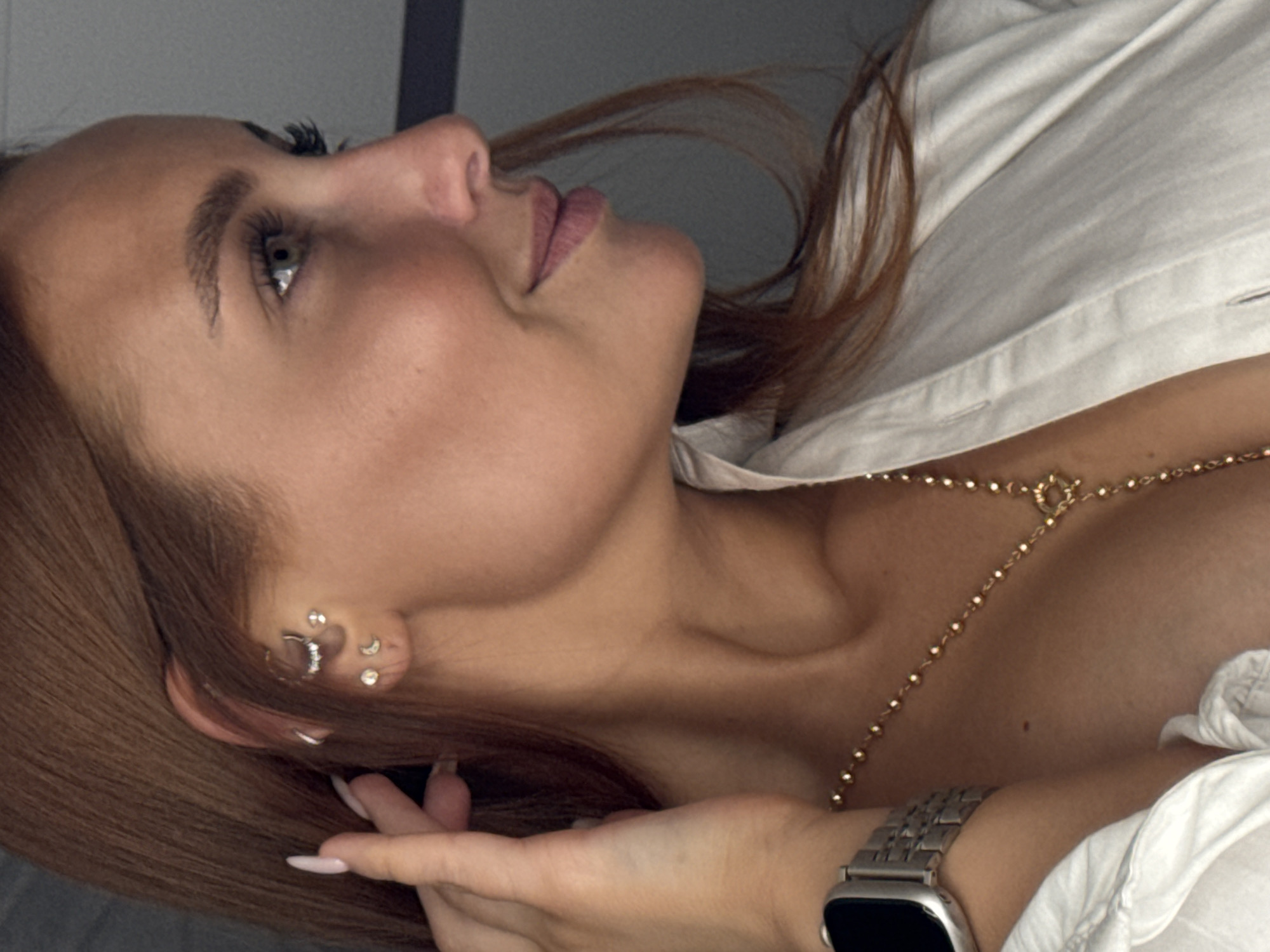A woman with fair skin and brown hair is lying down, looking upwards and to the left. She is wearing a white shirt and a gold body chain. A silver smartwatch is visible on her wrist. Her left ear has multiple piercings. No evidence suggests the image was taken in Chaikovsky, Russia.