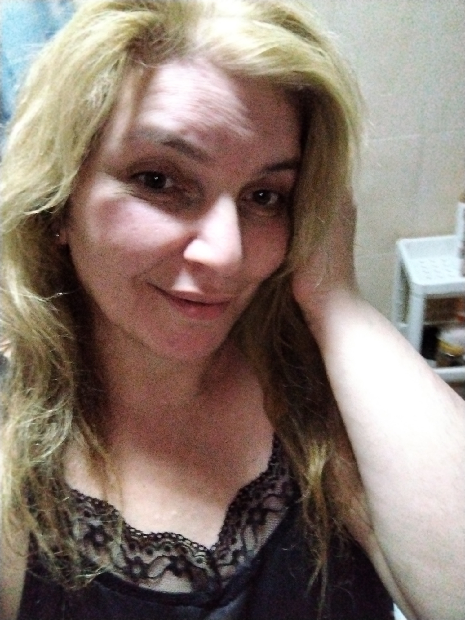 A mid-shot of a fair-skinned woman with blonde, wavy hair. She is wearing a dark top with lace trim. She appears to be indoors. A white plastic shelving unit is visible in the background. The location is not discernible as Al Mas'oudi, United Arab Emirates without further information.
