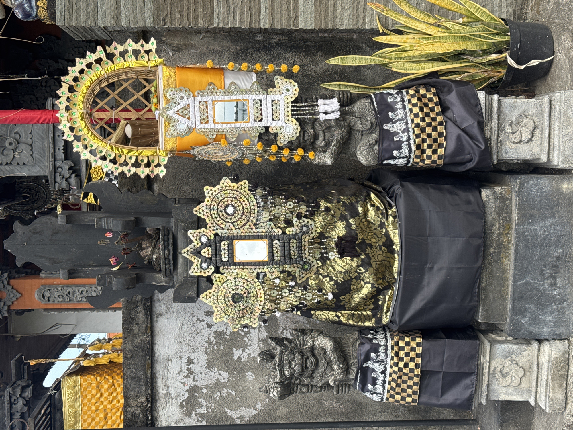 The image depicts an outdoor shrine in Sukawati, Indonesia, likely in front of a building or temple. The shrine features two stone statues of mythical creatures, possibly guardians, flanking a central altar. These statues are adorned with black and gold patterned cloths and black fabric.
The altar itself is elaborately decorated with ornate golden headdresses, mirrored embellishments, and hanging yellow tassels. A black fabric with a floral pattern covers the base of the central altar. Above the left statue, a large golden fabric with a checkered pattern is visible, suggesting it is part of the shrine's larger structure.
To the right of the shrine, a potted snake plant with long, variegated leaves adds a touch of greenery to the scene. The background consists of a textured stone wall and architectural elements that suggest a traditional Indonesian style. The overall impression is one of cultural and religious significance, with meticulous attention to detail in the adornment of the shrine.