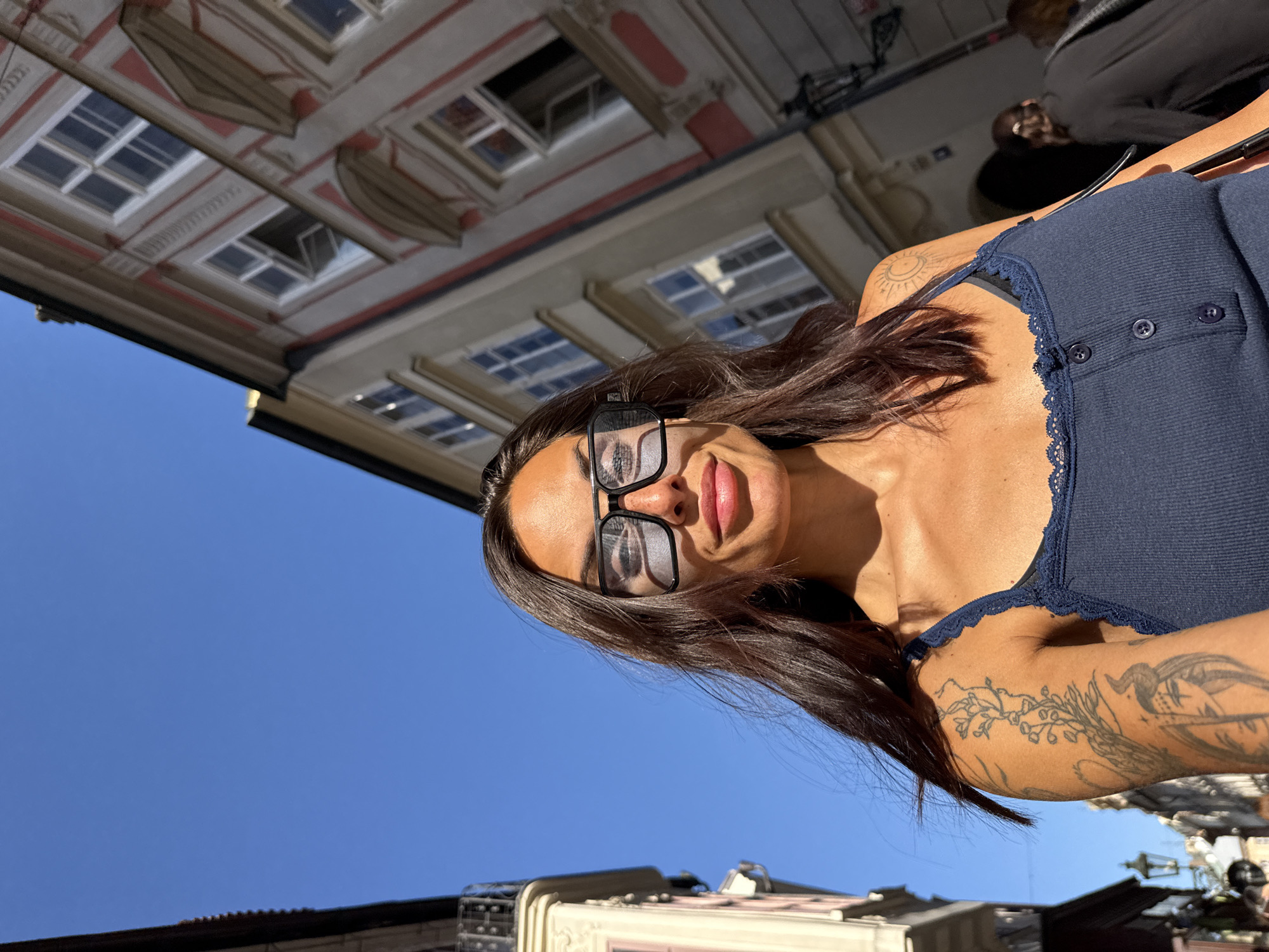 A woman is taking a selfie on a street in Prague, Czechia. She has long, dark hair and is wearing large, black-rimmed sunglasses and a navy blue tank top with lace trim. She has tattoos on her left arm. In the background, there are old European buildings with detailed architecture and arched windows. The sky is clear and blue.
