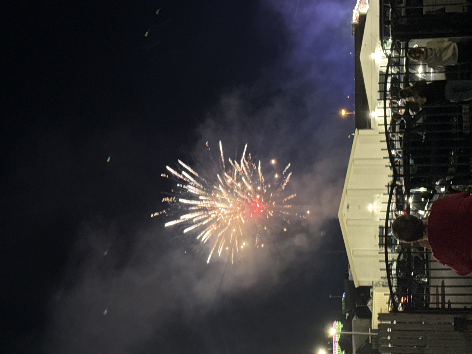The image depicts a nighttime scene featuring a fireworks display over Tramore, Ireland. In the foreground, a person wearing a red shirt is visible from the back, standing behind a black metal fence. Further back, two other individuals, one with long blonde hair and the other with blonde hair, are also standing behind the fence, looking towards the fireworks.
The middle ground of the image is dominated by a large, white building with illuminated windows. Several cars are parked in front of the building and behind the fence.
The sky is dark and filled with smoke from the fireworks. A large firework is exploding in the upper left quadrant of the image, emitting bright white and yellow sparks, with a prominent red center. Other smaller sparks and trails of light are scattered throughout the sky. There are also some faint green lights visible in the upper portions of the sky, the origin of which is unclear. The overall atmosphere suggests a public event or celebration taking place in the town.