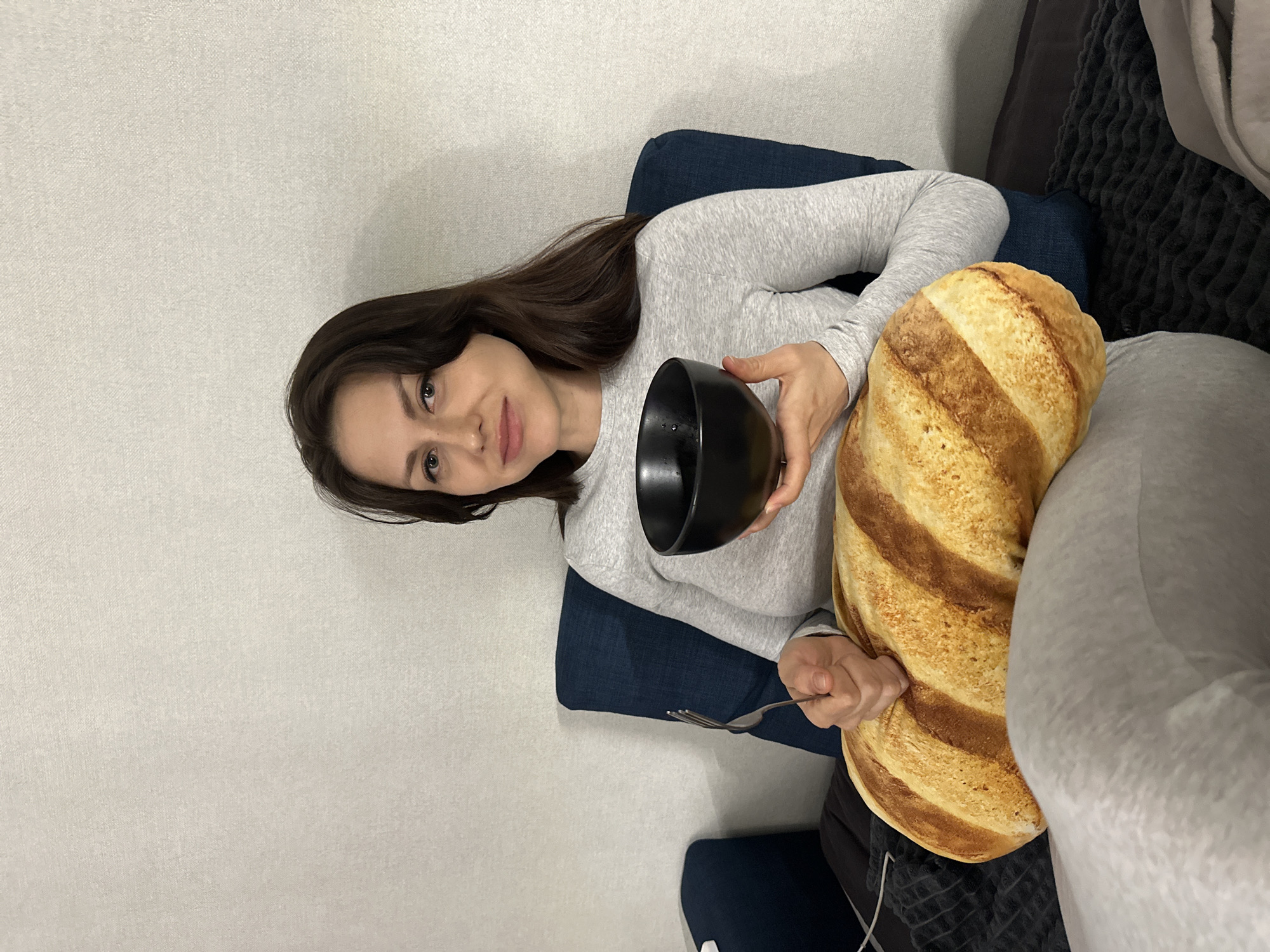 A woman is seated indoors in Murino, Russia. She is holding a black bowl in one hand and a fork in the other, and there is a pillow shaped like a loaf of bread on her lap. The woman has long dark hair and is wearing a gray long-sleeved shirt and gray sweatpants. She is sitting on a dark blue chair next to a gray and dark gray sofa. The background is a light-colored wall.