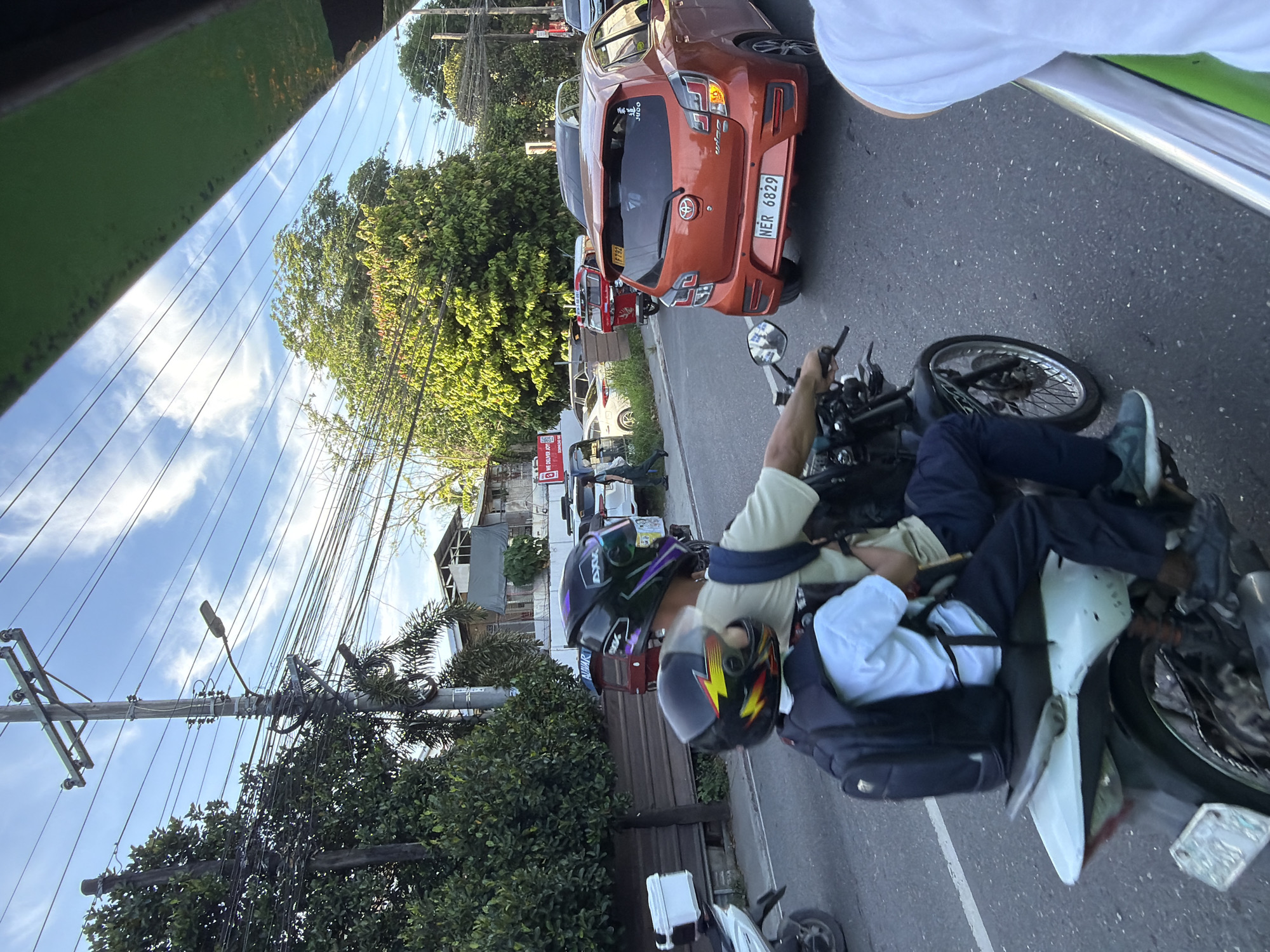 The image shows a street scene in Davao City, Philippines. The focal point is a motorcycle with two occupants. The driver wears a black helmet with purple and green accents, and a light-colored t-shirt. The passenger, a child, wears a black helmet with yellow and red lightning bolt designs and a white t-shirt. Both are wearing backpacks. They are riding on a paved road with other vehicles present, including a bright orange car with the license plate "NER 6829" and a red tricycle. In the background, there are trees, utility poles with numerous wires, and buildings. The sky is blue with scattered white clouds. The overall scene depicts a typical urban traffic environment.