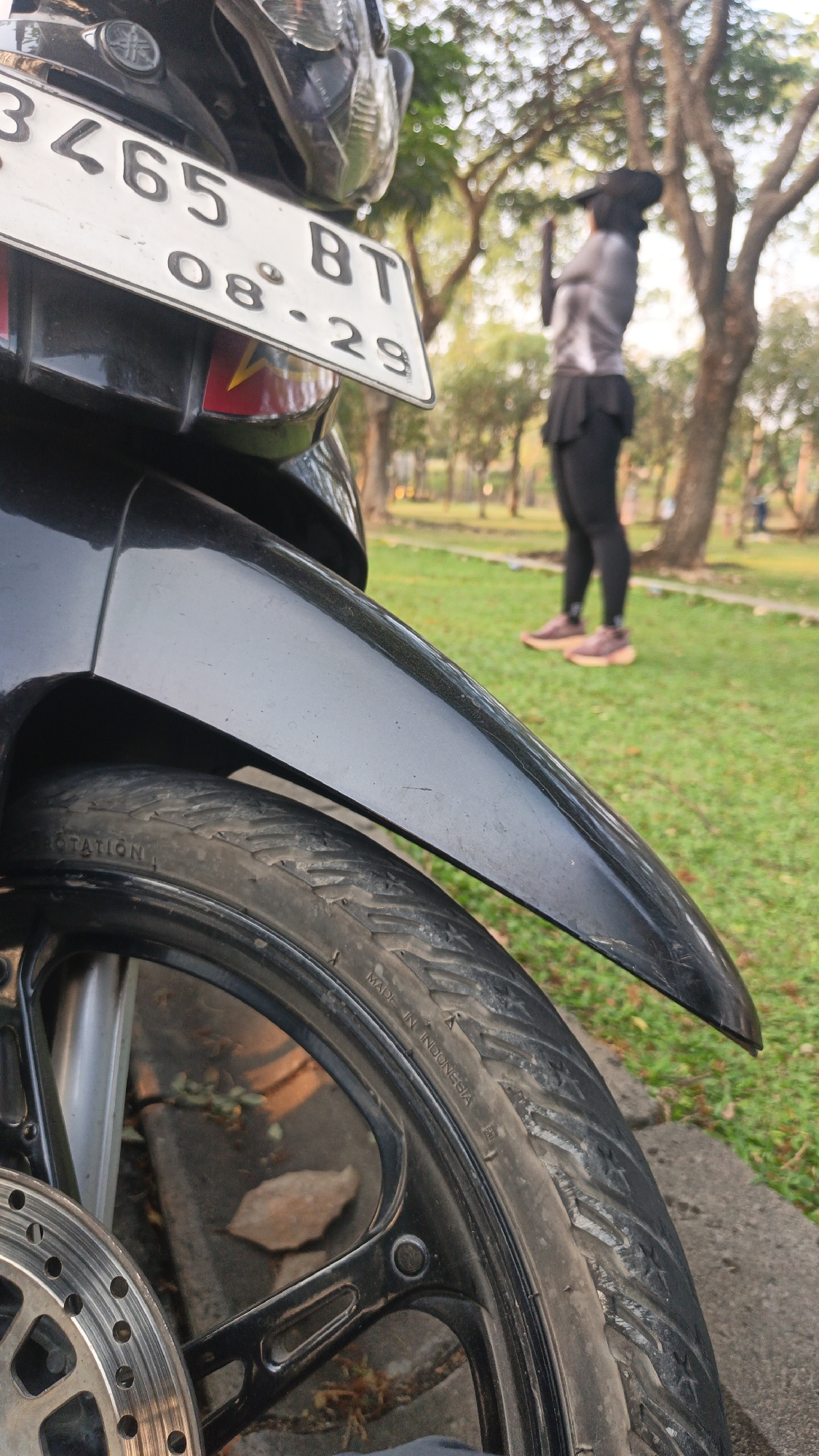 An outdoor, low-angle, full shot captures a close-up of a black motorcycle fender and front wheel with a license plate in the upper left quadrant. The license plate reads "3465 BT 08-29". In the background, a person wearing a black head covering, a gray and black long-sleeved shirt, black leggings, and pink athletic shoes is standing in a grassy park area with trees. The person appears to be looking at their phone. The environment suggests a park in Bacin, Indonesia, with sunlight filtering through the trees.