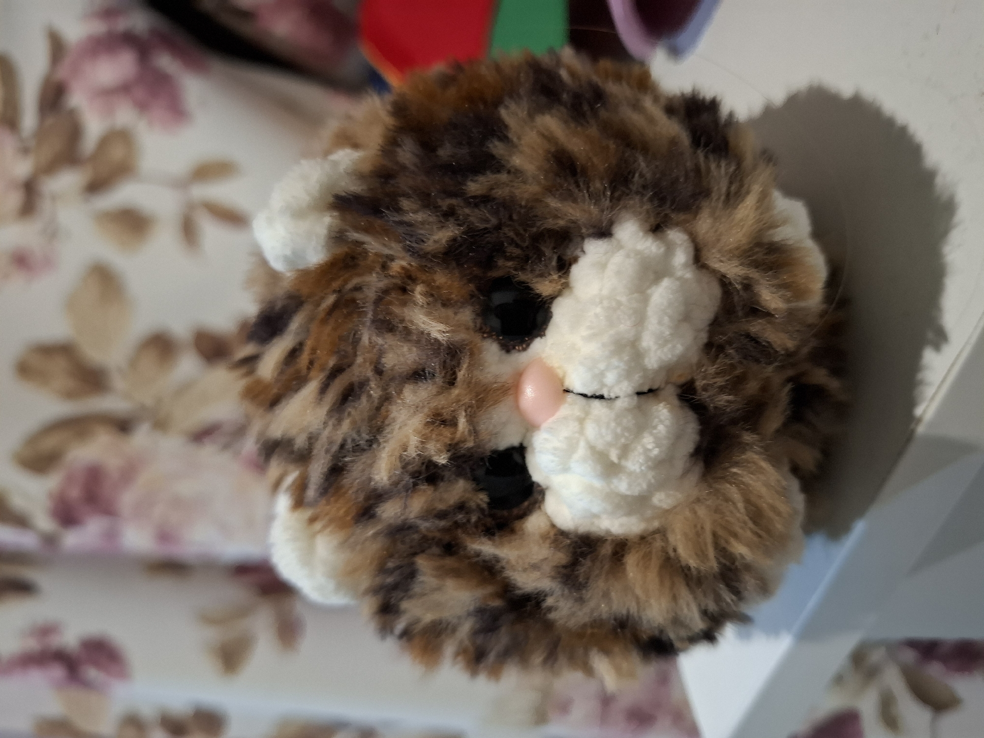 A close-up, front-facing shot of a fluffy, brown, and black striped yarn pom-pom cat toy is positioned on a white surface. The toy has prominent white yarn features for its ears, muzzle, and cheeks. Its eyes are black, and its nose is a small, pink bead. The background is softly blurred, showing a floral curtain with pink and beige patterns. To the right of the toy, a portion of a red object and a green object are visible, out of focus. The toy casts a shadow to its right on the white surface. This image was taken on March 2, 2024, and there is no information available to determine its location in Korenovsk, Russia, as it is a close-up of a toy without any identifying contextual elements of that specific city.