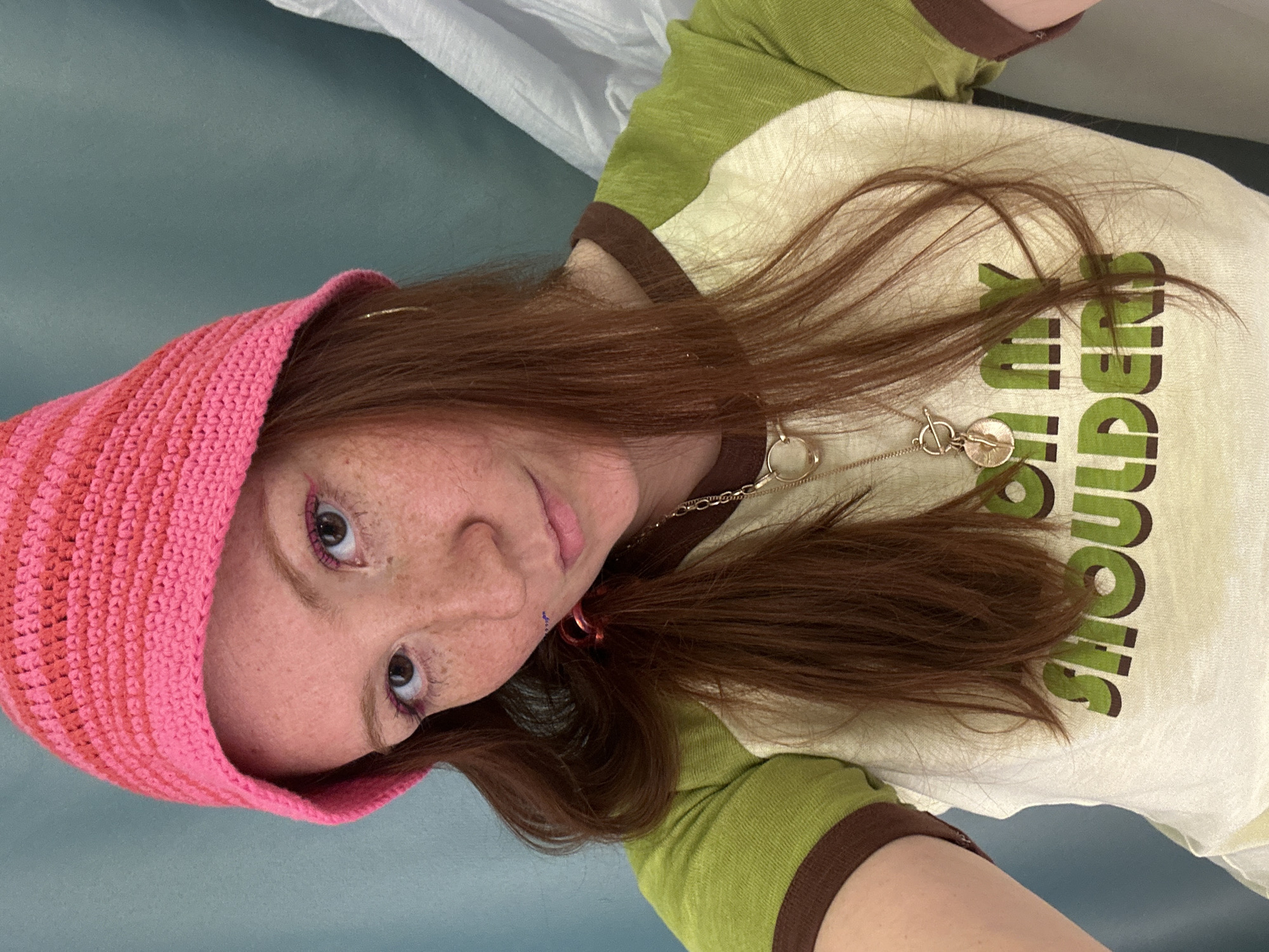 A woman with red hair, fair skin, and freckles is facing the camera. She is wearing a bright pink knitted hat and has pink eyeliner on her upper eyelids. She has a piercing on the left side of her lower lip. She is wearing a two-toned shirt with light green sleeves, a brown collar, and a light beige body with dark green letters. She has a gold necklace with a circular pendant. The background appears to be a light gray or blue surface, possibly a wall or floor. A white fabric is visible in the top portion of the image. There is no indication of the location being in Nizhny Novgorod, Russia.
