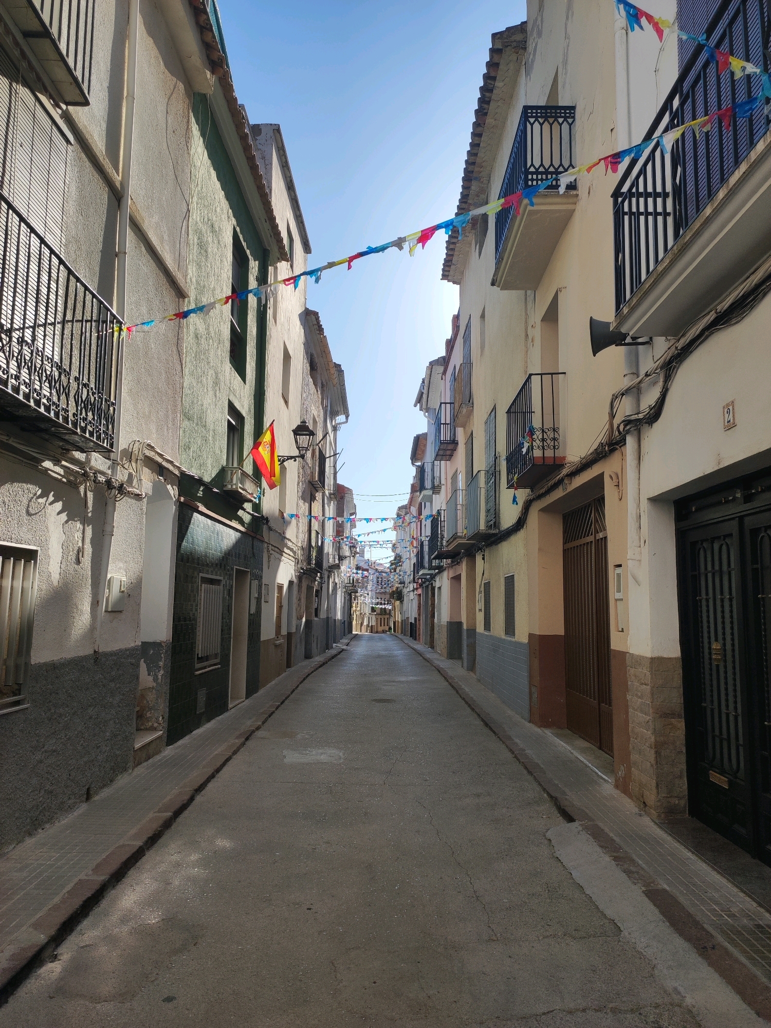 The image depicts a narrow, European-style street in the city of Sueras, Spain. The street is paved with asphalt, with a cobblestone sidewalk running along the left side. Buildings line both sides of the street, with balconies adorned with colorful bunting. A Spanish flag is visible hanging from one of the buildings. The sky is clear and blue, suggesting a sunny day. There are no people visible in the image.
