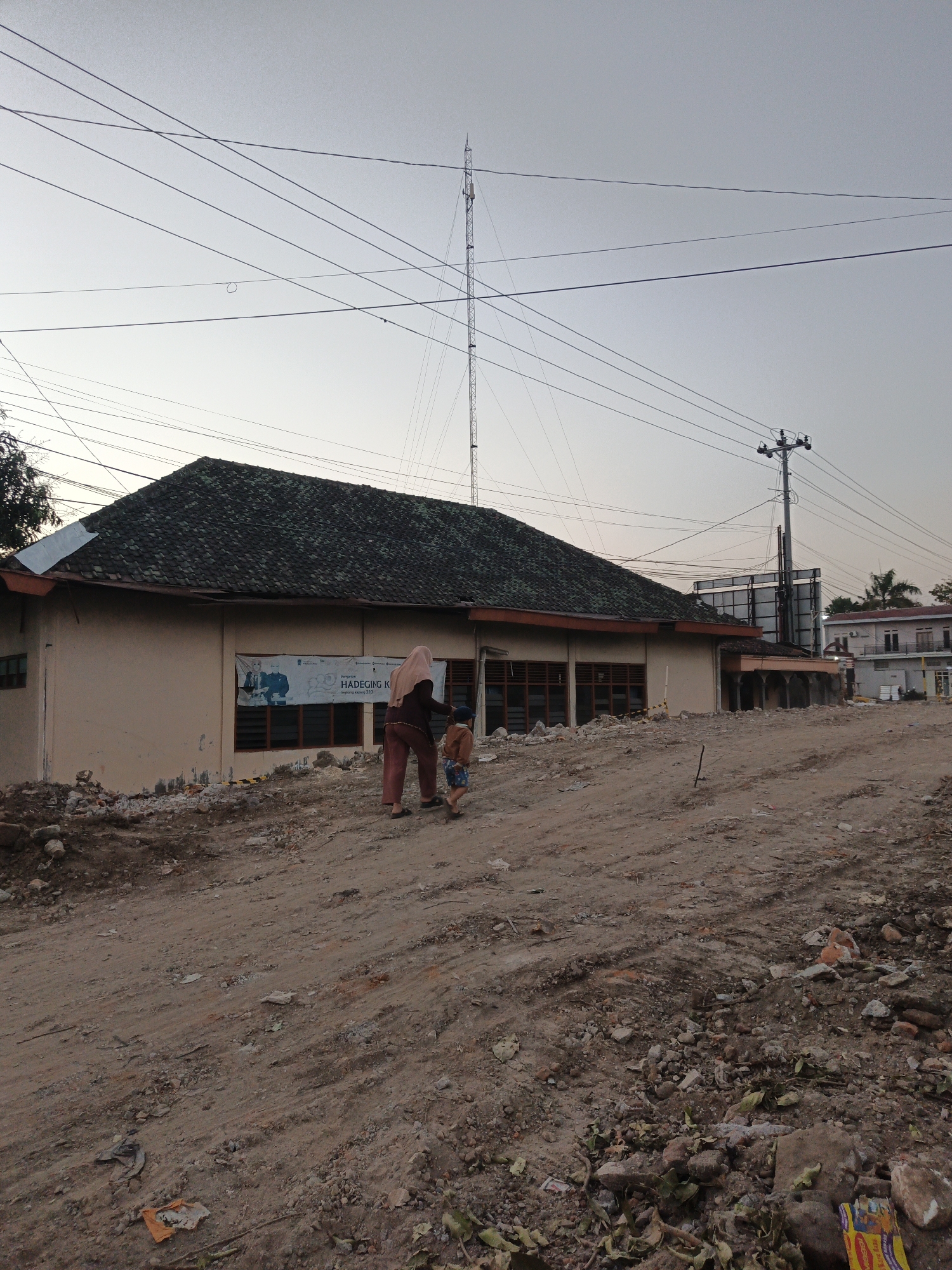 In Planggu, Indonesia, a person wearing a headscarf and brown clothing walks on a dirt path towards a building. The building has a dark green tiled roof and light brown walls. A banner with text and images is hanging on the building's exterior. A young child in a blue hat and blue shorts walks next to the person. The ground is uneven and covered with dirt, rocks, and debris, suggesting construction or demolition activity. In the background, utility poles with numerous wires stretch across the sky, and a tall tower, possibly a radio tower, is visible. The sky is a pale gray, indicating either dawn or dusk.