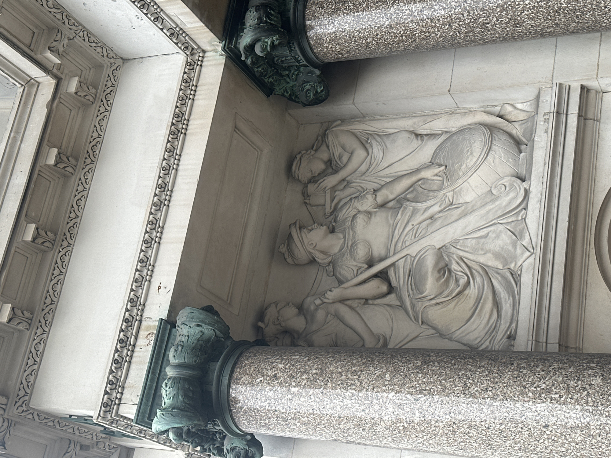 The image shows a section of the facade of a building, likely in London, United Kingdom, featuring a carved relief and ornate architectural elements. Two mottled grey stone columns with decorative bronze-green capitals bracket the relief. The relief depicts three classical figures, draped in flowing robes, in a reclining position. One figure is wearing a helmet and holding a long object, possibly a spear or staff. Other parts of the building are decorated with patterns.