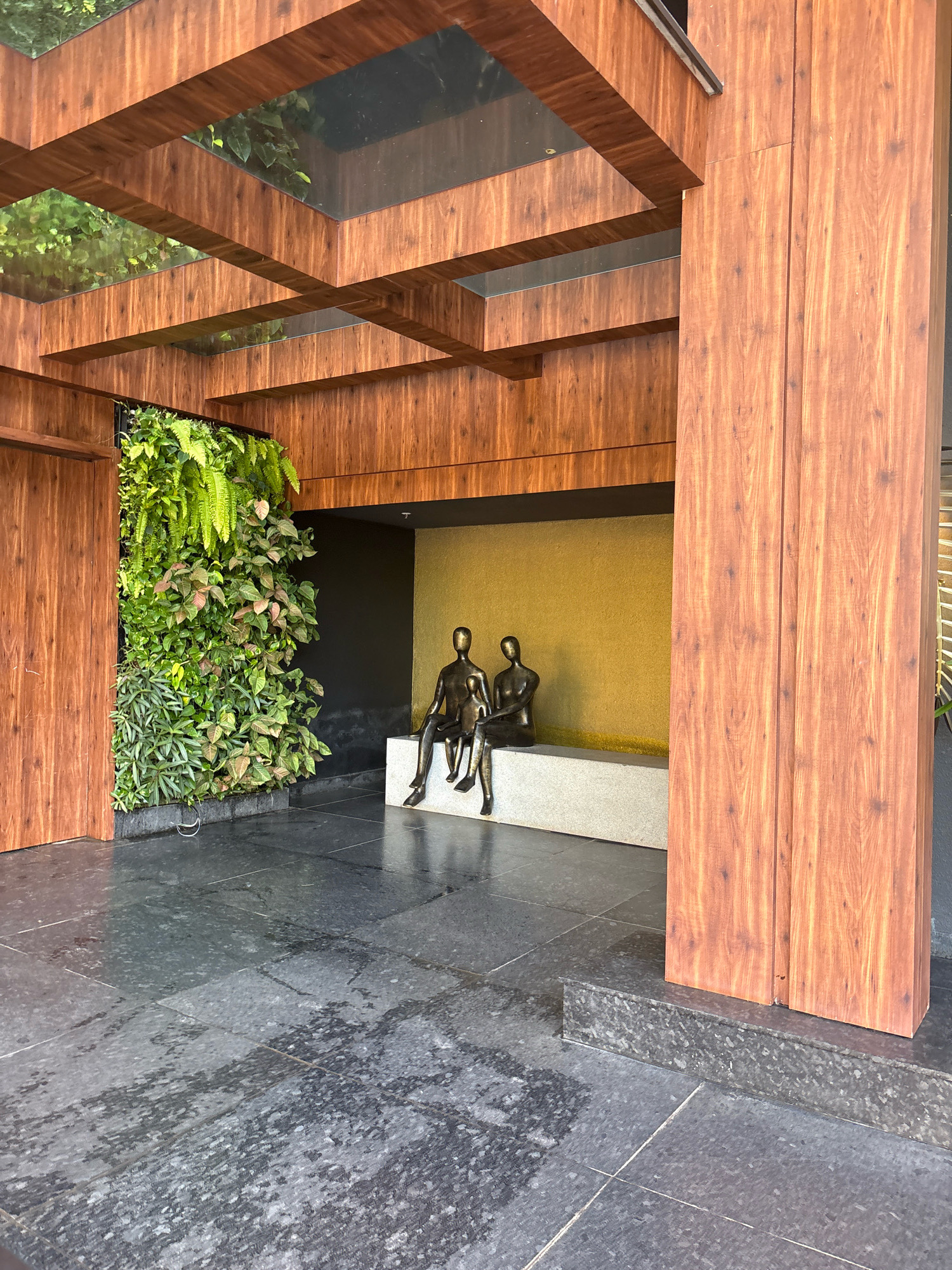 The image depicts the entrance area of a modern establishment, likely a hotel or a restaurant, in Vagator, India. The architecture features extensive use of wood paneling, both for the structural elements like the large pillar on the right and the overhead beams, as well as for the door on the left. Above, a wooden lattice structure with glass panels allows natural light to filter through, revealing glimpses of greenery.
Dominating the left side of the frame is a lush vertical garden, a wall densely covered with various green plants, creating a vibrant contrast with the surrounding wooden and stone surfaces. In the center, a seating area is established with a light-colored stone bench against a backdrop of a textured golden-yellow wall. Two bronze-toned abstract human figures are seated on the bench, appearing to be in conversation or contemplation. The ground is paved with large, dark grey, textured stone tiles, which appear damp in places, suggesting recent rain or cleaning. The overall impression is one of contemporary design blending natural elements with art.