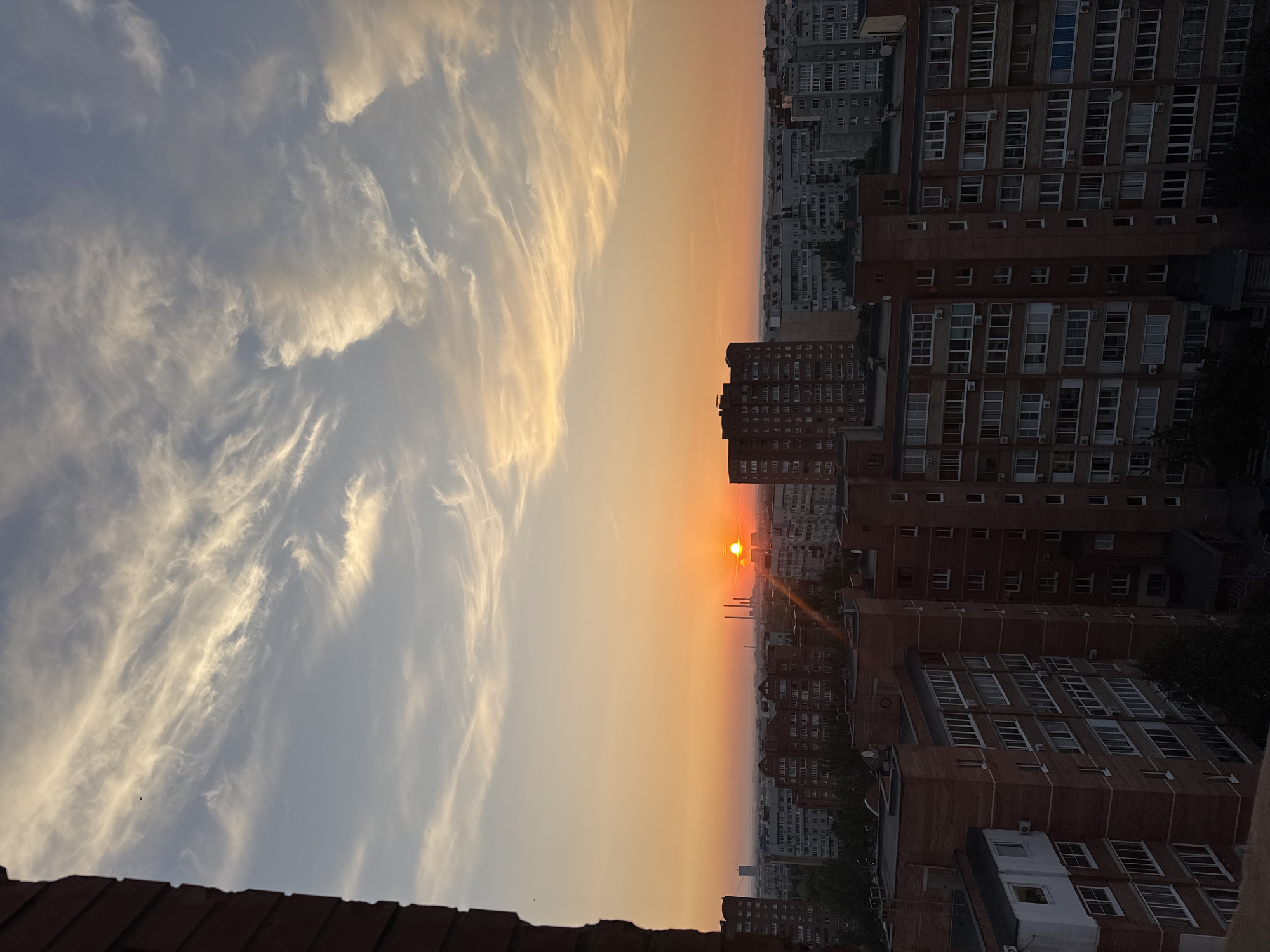 The image captures a panoramic view of a cityscape at sunset. The sun, a bright orange orb, is positioned low on the horizon, casting a warm glow across the sky and illuminating a cluster of tall buildings in the distance. Wispy, white clouds streak across the sky, adding texture and depth to the scene. In the foreground, the dark silhouette of a brick building juts into the frame from the left, providing a stark contrast to the soft hues of the sunset. The mid-ground is dominated by rows of apartment buildings, their windows reflecting the fading light. The architecture suggests a dense urban environment, with structures of varying heights and designs. The overall impression is one of a peaceful evening descending upon the city.