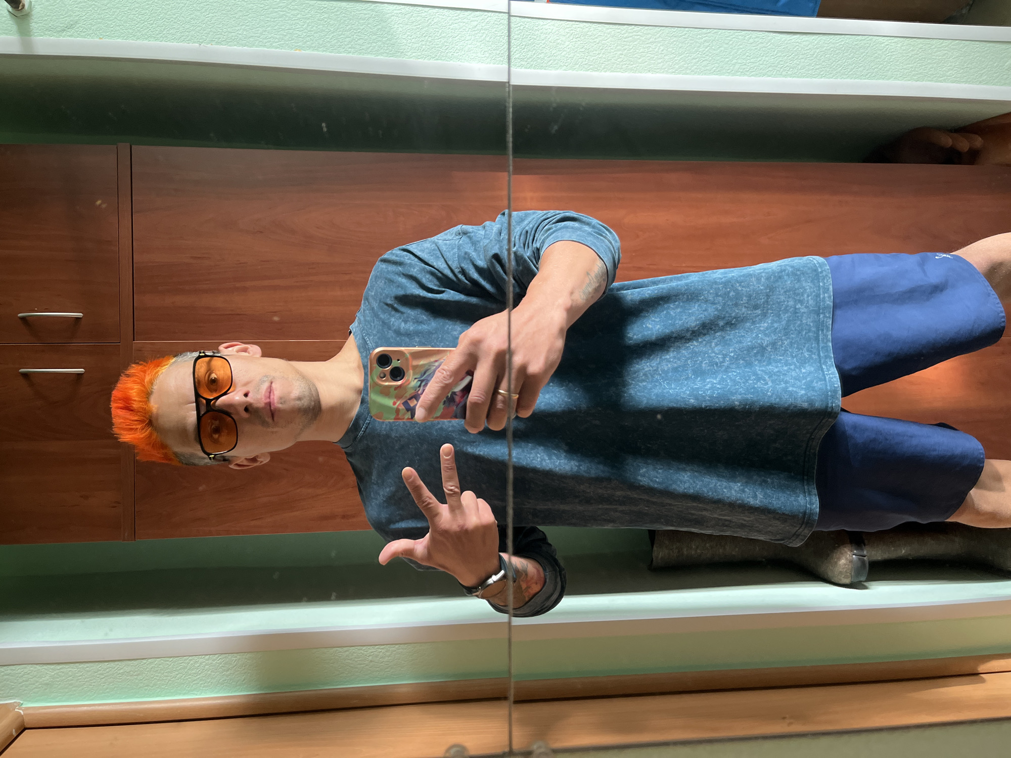 A man is standing in what appears to be a bathroom, taking a selfie in a mirror. He has bright orange hair, is wearing orange-tinted glasses, a blue-gray t-shirt, and blue shorts. He is holding a smartphone and making a peace sign with his hand. A dark wood cabinet is behind him and there is what looks like a person lying down under the cabinet. The location is possibly in a building in Chaikovsky, Russia.