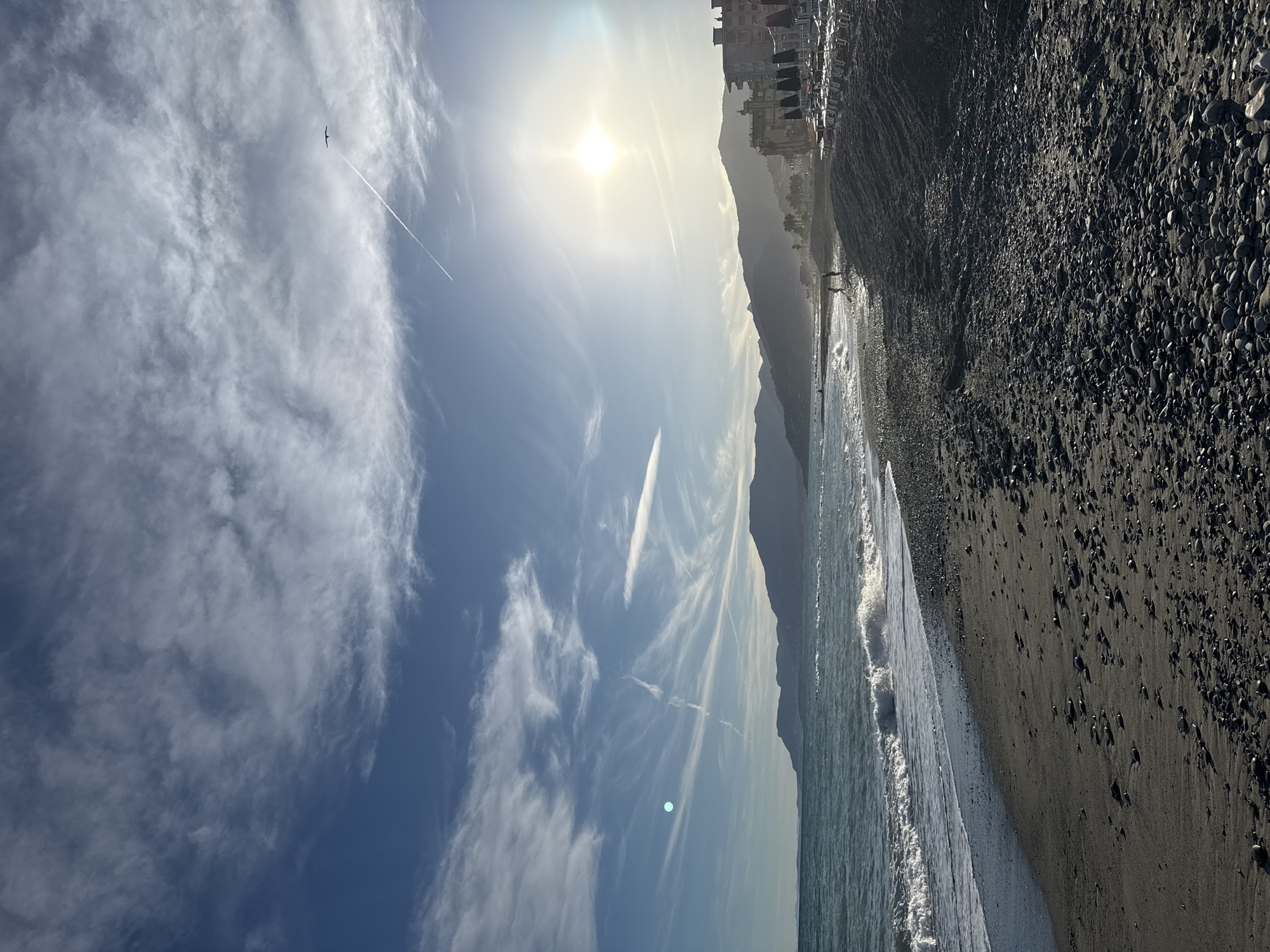 This is a wide-angle, vertical shot of a beach in Vallecrosia al Mare, Italy, with the Ligurian Sea on the left and buildings and mountains on the right. The sky is a vibrant blue with wispy white clouds and a bright sun creating a lens flare. A plane's contrail stretches across the sky, and a bird is silhouetted against it. The sea has small waves breaking on the shore. The beach itself is composed of dark sand and pebbles, with a line of beach chairs and umbrellas set up near the buildings. Two people are visible walking on the beach in the distance, and another person appears to be standing near the water. The background features rolling hills or mountains, some of which are covered in vegetation. The scene is bathed in sunlight, suggesting it is daytime.