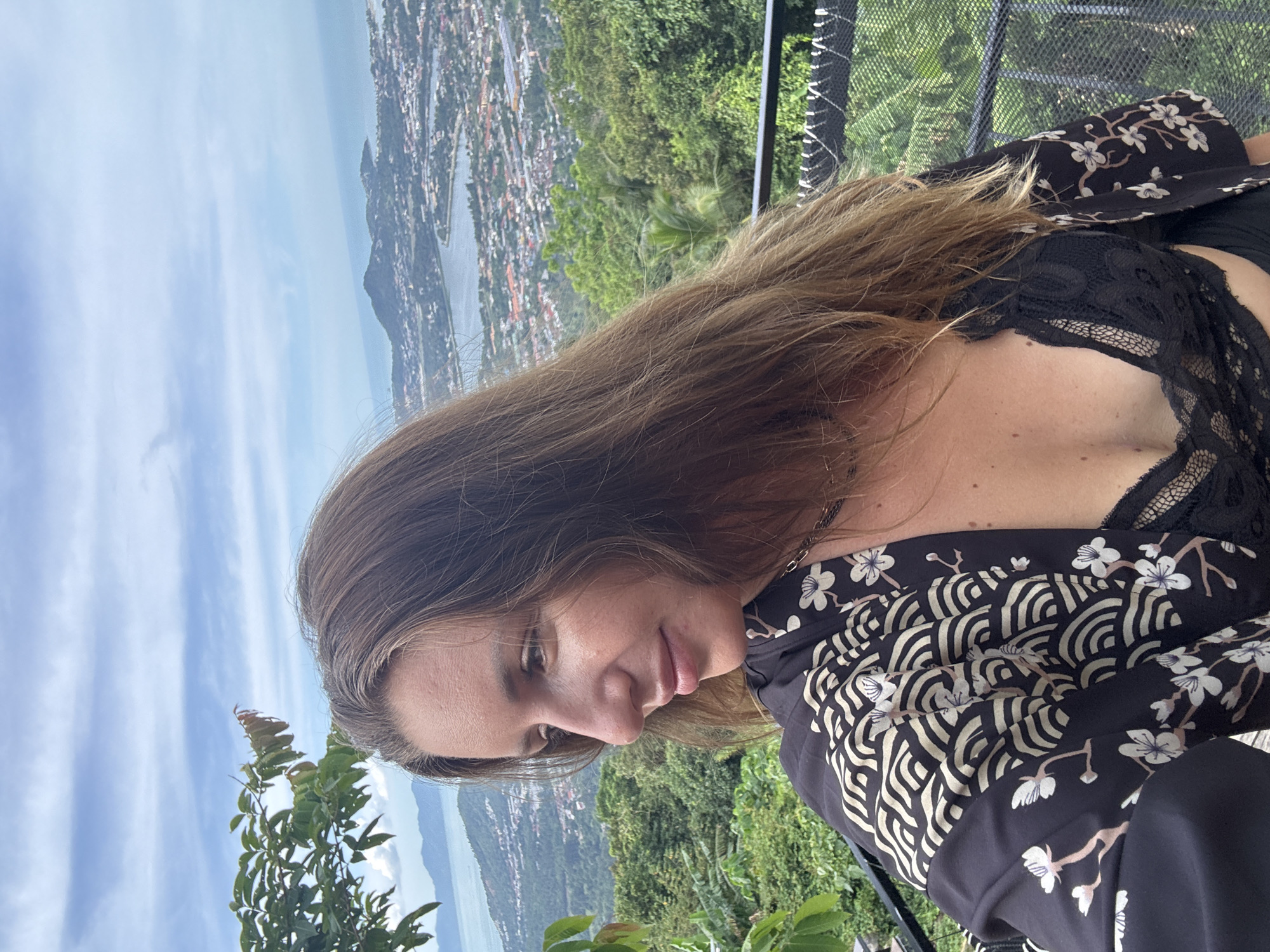 A woman with long, brown hair and fair skin is pictured from the chest up, looking to the left of the frame. She is wearing a black, floral-patterned kimono over a black lace bralette. In the background, a scenic view of a coastal city unfolds, with buildings nestled amongst lush green hills that slope down to the ocean under a cloudy sky. The scene appears to be taken from an elevated viewpoint, possibly a balcony or overlook, as indicated by a dark railing in the foreground.