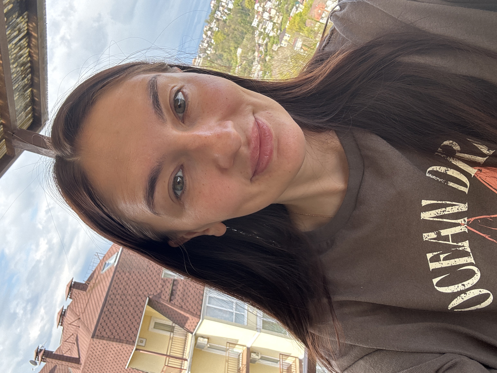 A selfie of a fair-skinned woman with dark brown hair and eyes is taken outdoors. The woman is wearing a brown t-shirt with white lettering that reads "OCEAN DR". In the background, there is a cityscape with trees and buildings. The location is likely in Sochi, Russia, given the architectural style of the building in the foreground and the hilly terrain.