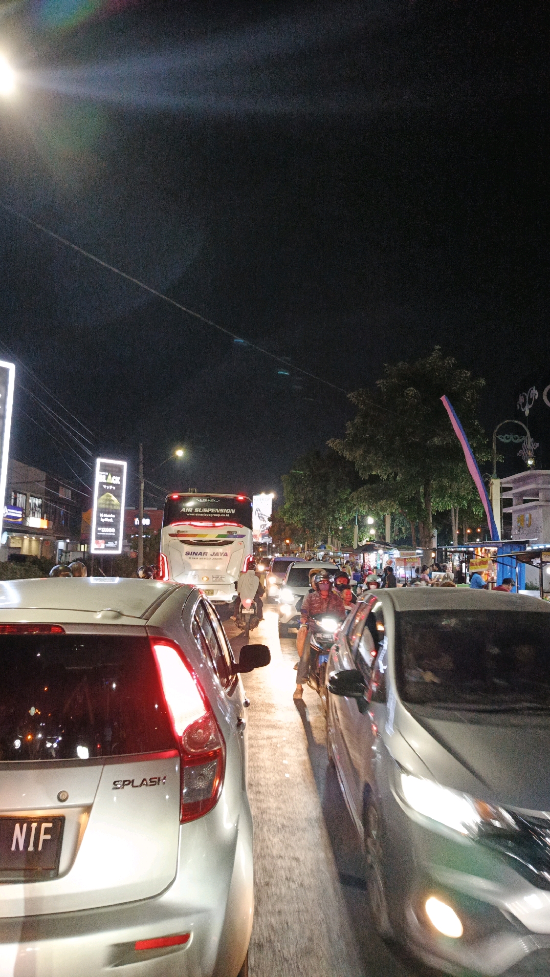 The image depicts a nighttime traffic scene in Surakarta, Indonesia, characterized by heavy congestion. Several vehicles, including cars and motorcycles, are visible, suggesting a busy urban environment. A large bus with "SINAR JAYA" branding is prominent in the background, indicating it is likely part of a public transportation service. The presence of streetlights, illuminated signs on buildings, and vehicle headlights contributes to the nighttime ambiance and illuminates the road. The traffic appears to be stationary or moving very slowly, a common occurrence in congested city streets. The overall scene conveys the typical atmosphere of a bustling city at night, with the flow of traffic and the presence of commercial establishments.
