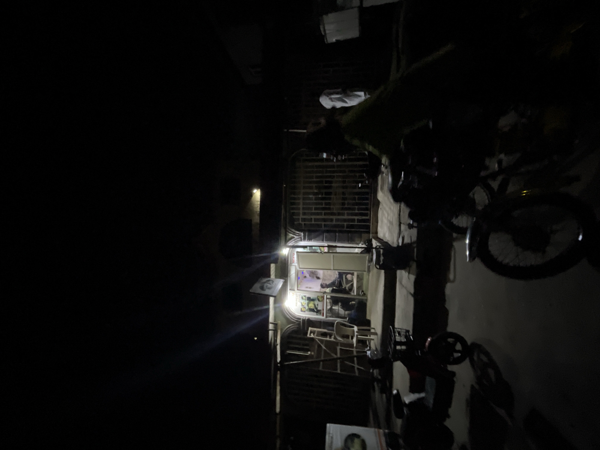 The image depicts a dimly lit street scene in Ado, Nigeria, at night. A bicycle is parked on the right. A building facade with a lit storefront labeled "NEXT HAIR CUT" is visible. A partially visible sign with a face on it is on the lower left. An adjacent building is barred with security grates. The light is minimal, casting deep shadows. The environment appears urban.