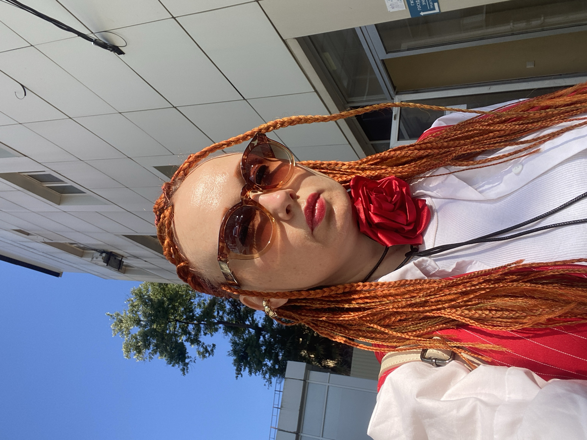 A person with bright orange braided hair and red lipstick stands in front of a modern building with a white facade, likely in Sochi, Russia, as indicated by the presence of a street sign with Russian text. The person is wearing large, tinted sunglasses, a white shirt, a red vest with white pinstripes, and a black necklace with a large red rose accessory. A white strap from a bag is visible over the shoulder. The background shows a clear blue sky with a single green tree to the left of the frame. The building's exterior is composed of large, light-colored panels arranged diagonally, with several windows. The lighting suggests it is daytime, with sunlight casting shadows.