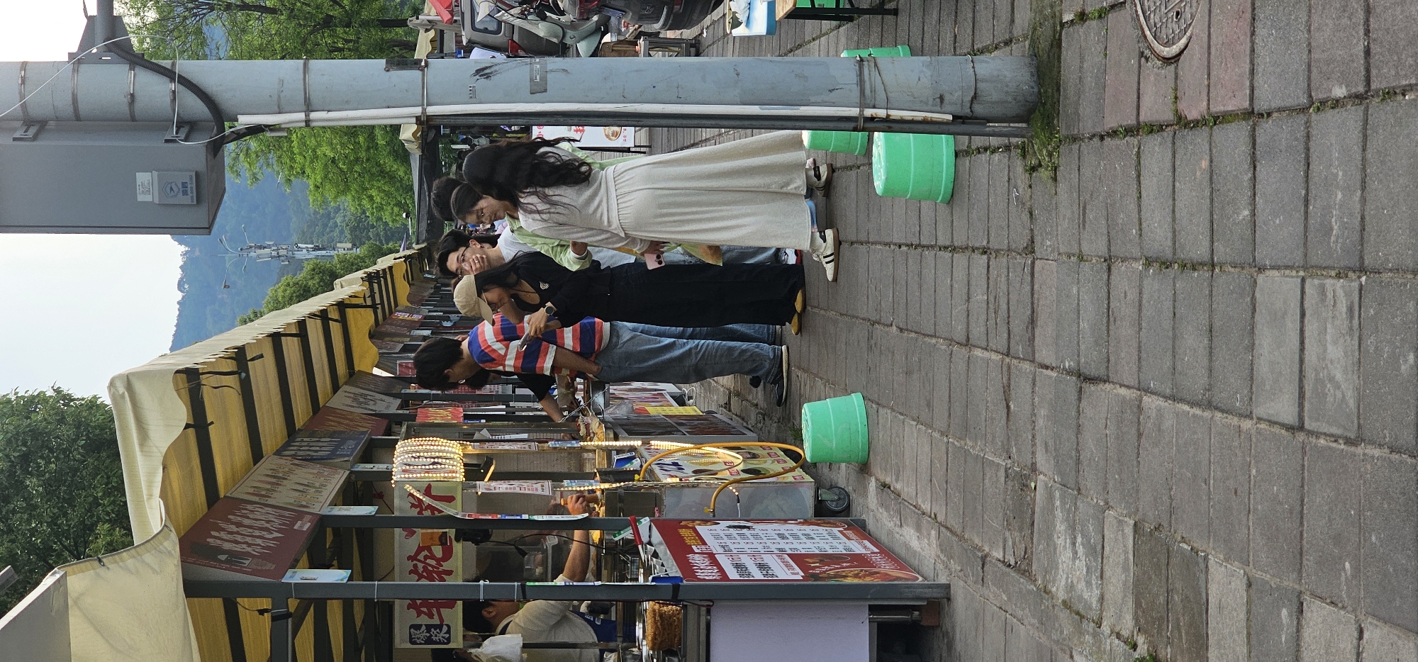 A street scene captured in Shapingba District, China, shows a row of street food vendors lined along a sidewalk. A group of people, consisting of at least four individuals, are standing at the vendors, presumably looking at the food offerings. One person, wearing a red and blue striped shirt, appears to be interacting with a vendor or selecting items. The vendor stalls are covered by a yellow and white striped awning and are displaying menus and items for sale.
Several green buckets are situated along the sidewalk. A metal utility pole with attached equipment runs along the side of the vendors. In the background, mountains and trees can be seen. The sidewalk is made of gray stone blocks.