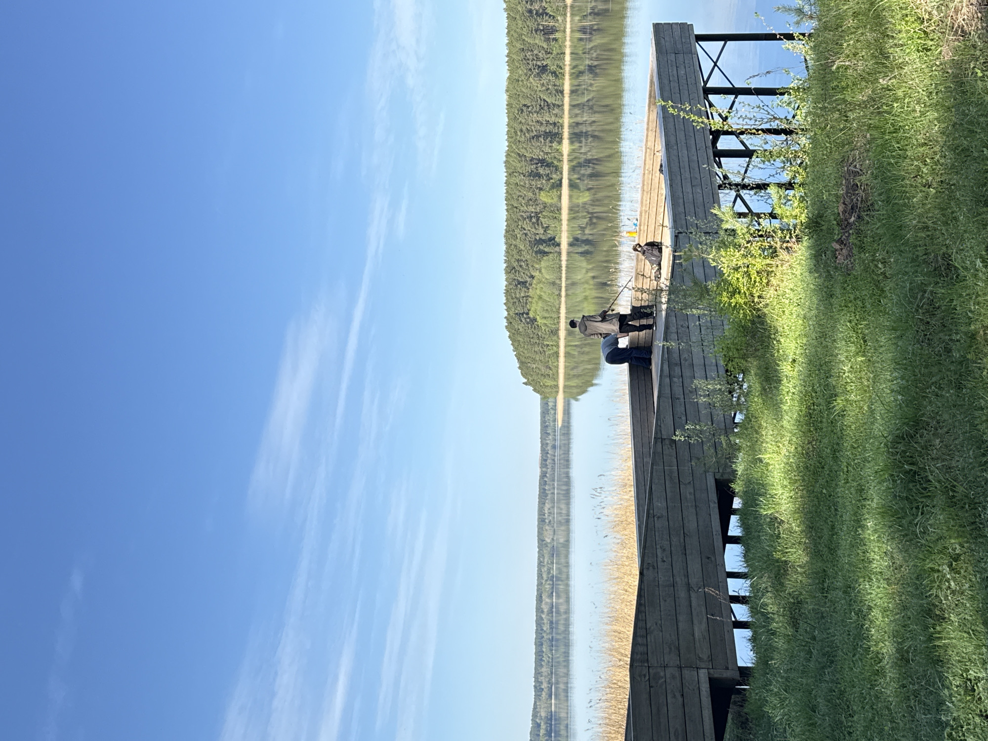 The image depicts a calm lake scene, likely during the daytime, featuring a wooden pier extending into the water. Two figures are on the pier, one potentially fishing, the other seated. The lake reflects the opposite shore, which is densely forested. The sky is blue with some wispy clouds. Grass grows along the edge of the pier. The specific location in Votkinsk, Russia, is not directly discernible from the image, though the presence of a forest-lined lake and wooden pier is consistent with the region's natural landscape.