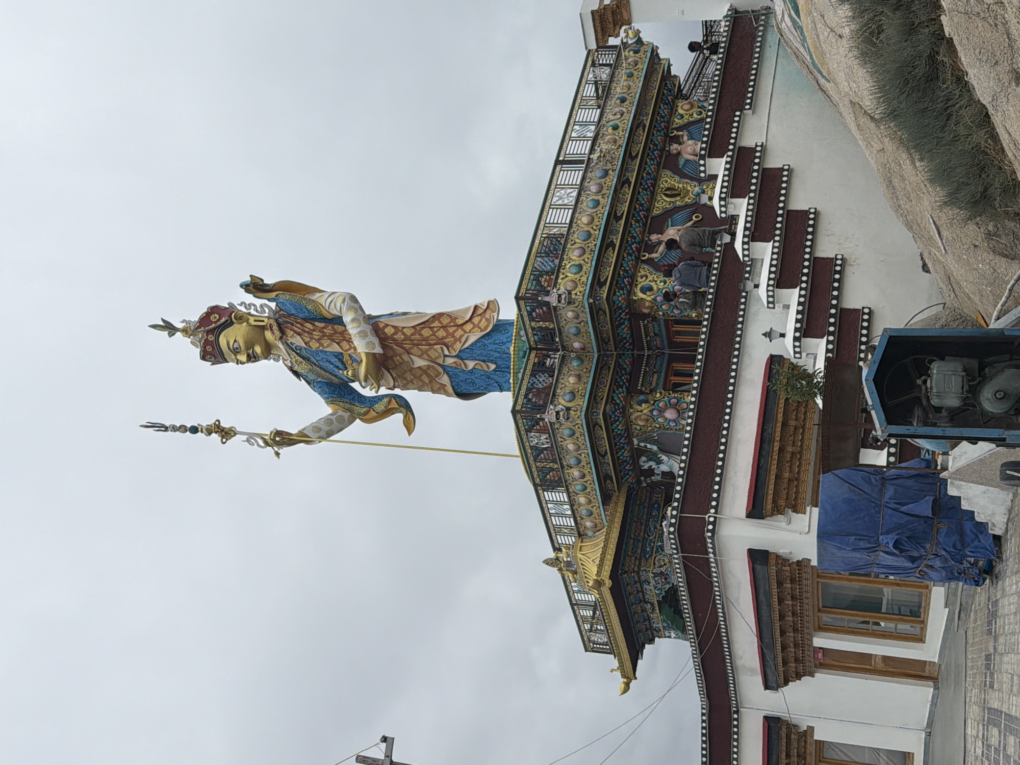 A large, golden statue of a deity, possibly a Buddhist figure, stands atop a multi-tiered, ornately decorated structure. The statue wears a red and gold crown and is clad in elaborate blue, gold, and red garments. It holds a staff topped with a trident-like ornament in its right hand and a vessel in its left. The structure beneath the statue features colorful murals depicting figures and intricate patterns. A white building with traditional wooden window frames and a dark red roof is visible below the decorated structure, with a blue tarp-covered object and a piece of machinery in the foreground. Several people are seen on the terraces and stairs of the structure. The sky is overcast, suggesting a cloudy day. The location is identified as Thiksey, India, likely referring to the Thiksey Monastery, famous for its large Maitreya Buddha statue.