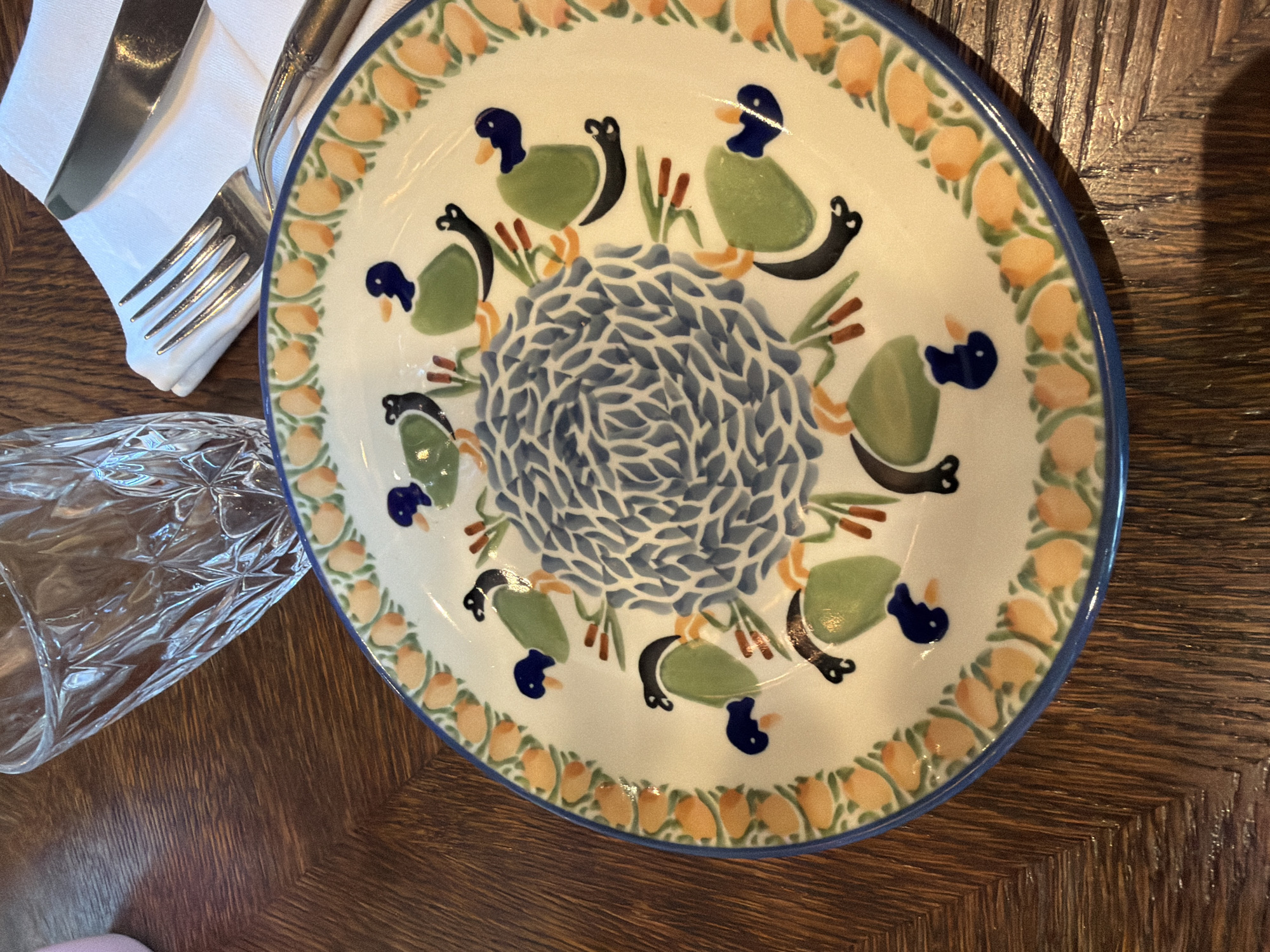 A close-up, high-angle shot captures a decorative plate with a duck motif placed on a wooden table. The plate features a central blue, swirled design surrounded by green ducks with blue heads and orange beaks. The outer rim of the plate is adorned with orange and green floral patterns. To the upper right of the plate, a white napkin holds a silver fork and knife. In the background, a clear glass with a faceted design is visible. The scene is set on a dark wooden table with a visible wood grain. The image does not provide any specific information about the location in Sabail Raion, Azerbaijan.