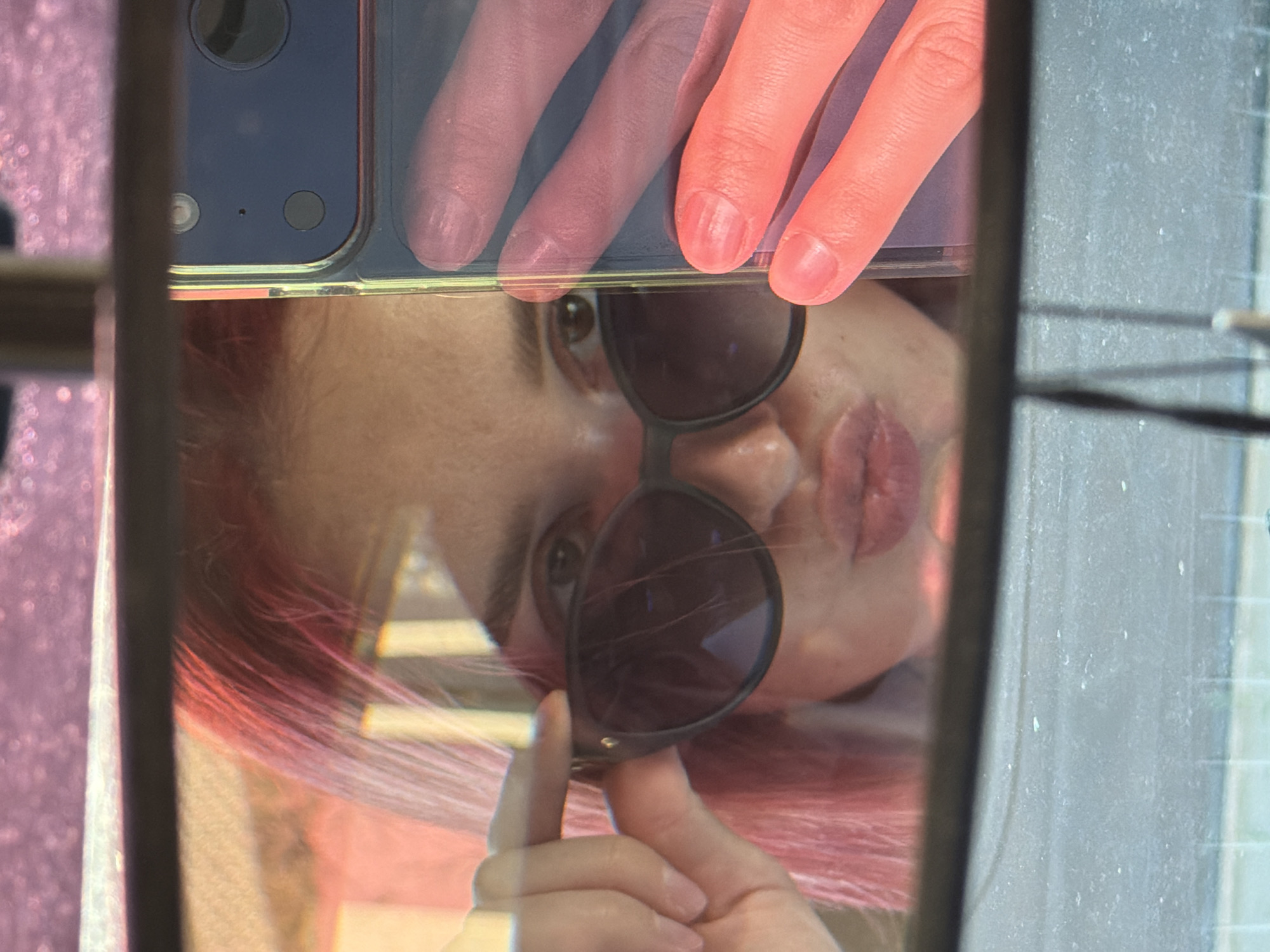 The image is a close-up selfie taken in a rearview mirror. A person with bright pink hair is holding the frame of dark sunglasses with their left hand, adjusting them near their eye. They are looking directly into the camera with their lips puckered. A smartphone is visible on the right side of the image, held by another hand with manicured fingernails. The background reflected in the mirror is blurred and appears to be an outdoor environment, possibly a street or a car interior. There are no discernible landmarks to identify a specific location within the city of Chaikovsky, Russia.