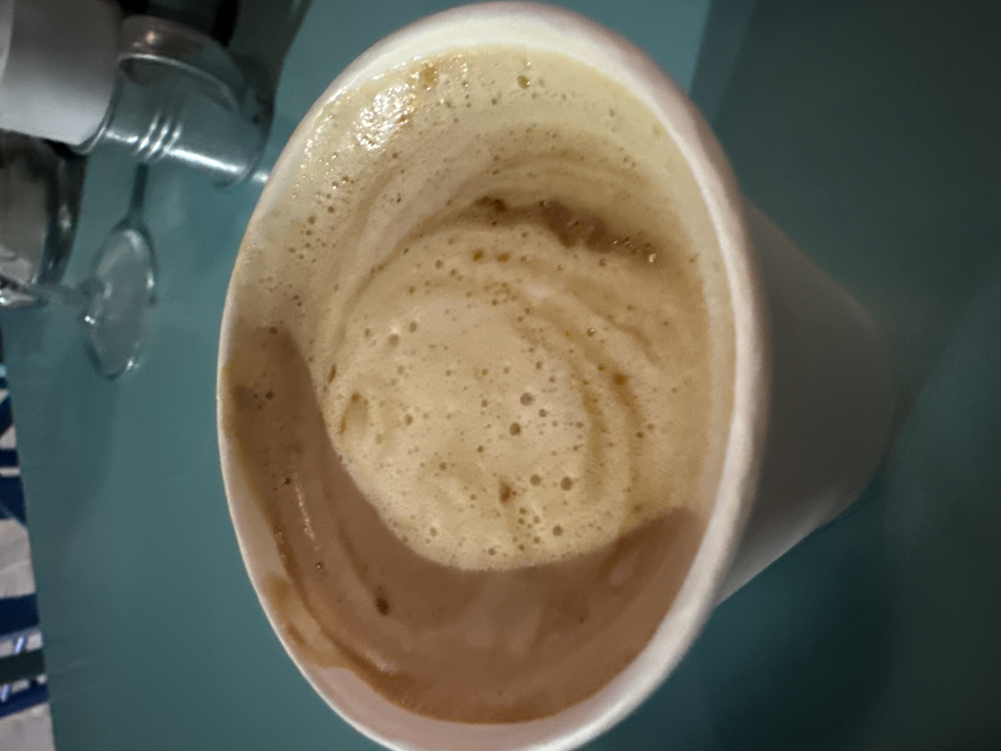 A close-up, overhead shot shows a white paper cup filled with a frothy coffee beverage. The coffee has a creamy, light brown foam with swirls of a darker brown, indicating it may be a latte or cappuccino. The cup is positioned on a teal-colored surface, likely a table. In the background, out of focus, are elements that suggest an outdoor or casual dining setting. These include a glimpse of a striped fabric in blue and white, a wine glass, and a small metal container holding white paper or napkins. The lighting suggests it is daytime, possibly outdoors given the reflections and color palette. The scene is devoid of any people or specific identifiable landmarks of Medinski Krš, Montenegro, beyond what can be inferred from the general setting.