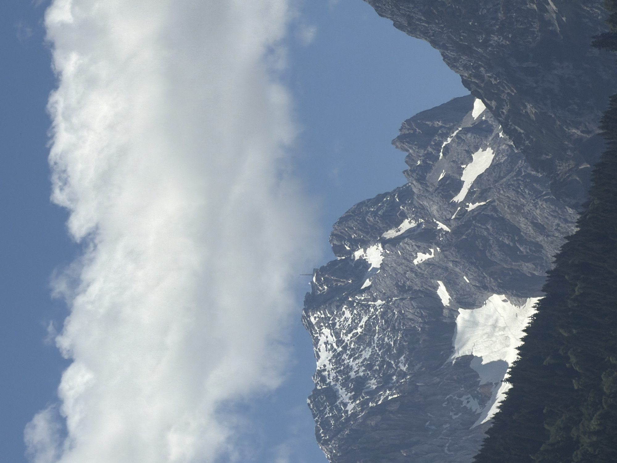 The image shows a mountainous landscape under a blue sky with scattered clouds. The foreground features a dense forest of dark green coniferous trees, suggesting a relatively high altitude or a northern climate. Behind the forest, rugged, grey mountain peaks rise sharply. Patches of snow are visible on the upper slopes and crevices of the mountains, indicating that the area is either at a high elevation or it is springtime with lingering snow. One prominent peak in the center has a structure resembling a telecommunications tower on its summit. The lighting suggests it is daytime, likely midday or afternoon, with the sun casting shadows that highlight the contours of the mountains. The overall scene is natural and untamed, with no visible signs of human structures other than the tower on the peak.
