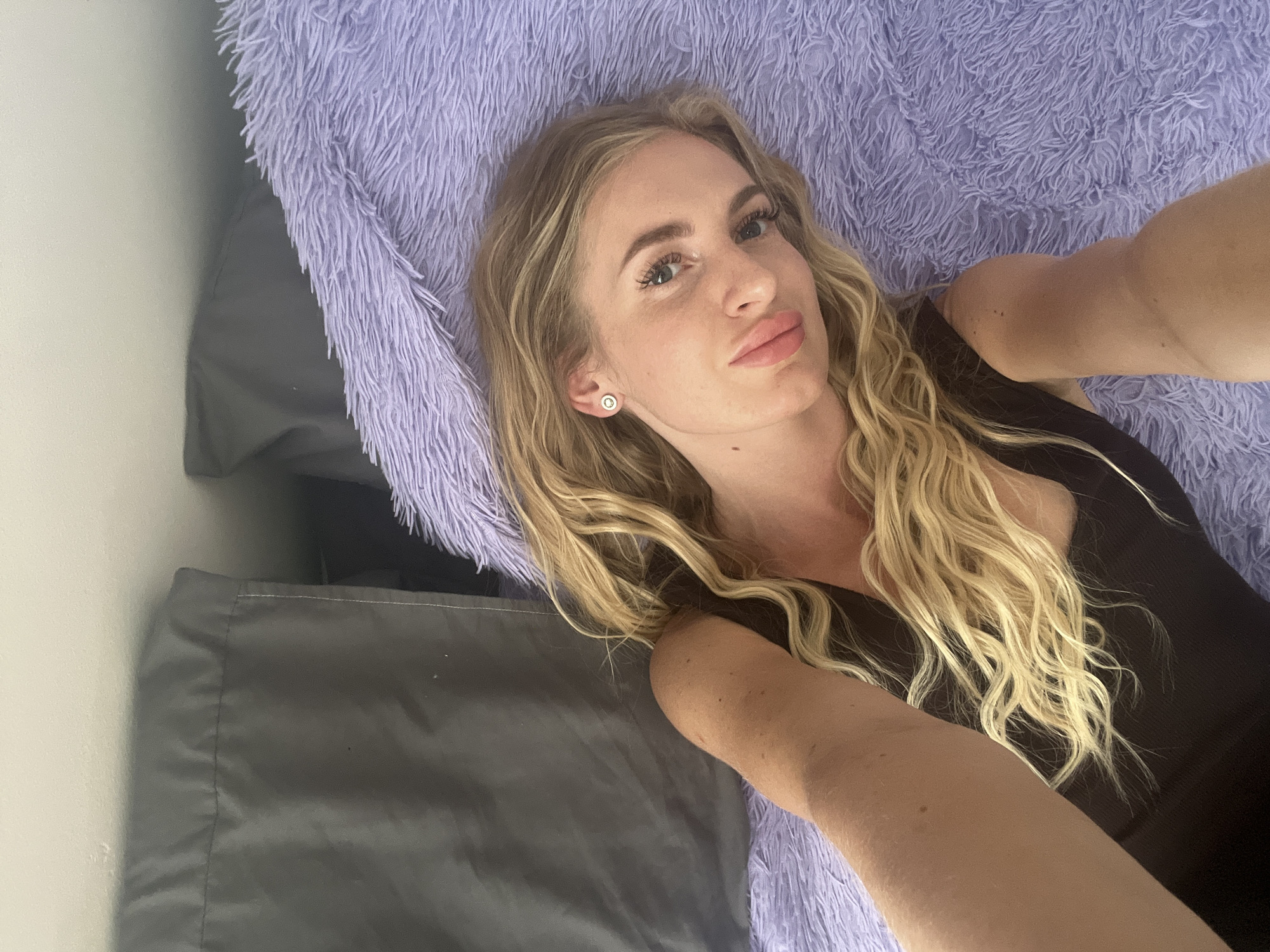 A blond woman with fair skin is lying on a bed. She has blond, wavy hair and is wearing a dark-colored top. The woman is angled to the left of the frame and looking into the camera with slightly pursed lips. Her head is resting on a purple fluffy blanket. Adjacent to the fluffy blanket, there is a gray pillow and bedsheet. The background includes an off-white wall. The location of the city of Budva, Montenegro, is not discernable from the provided visual data.