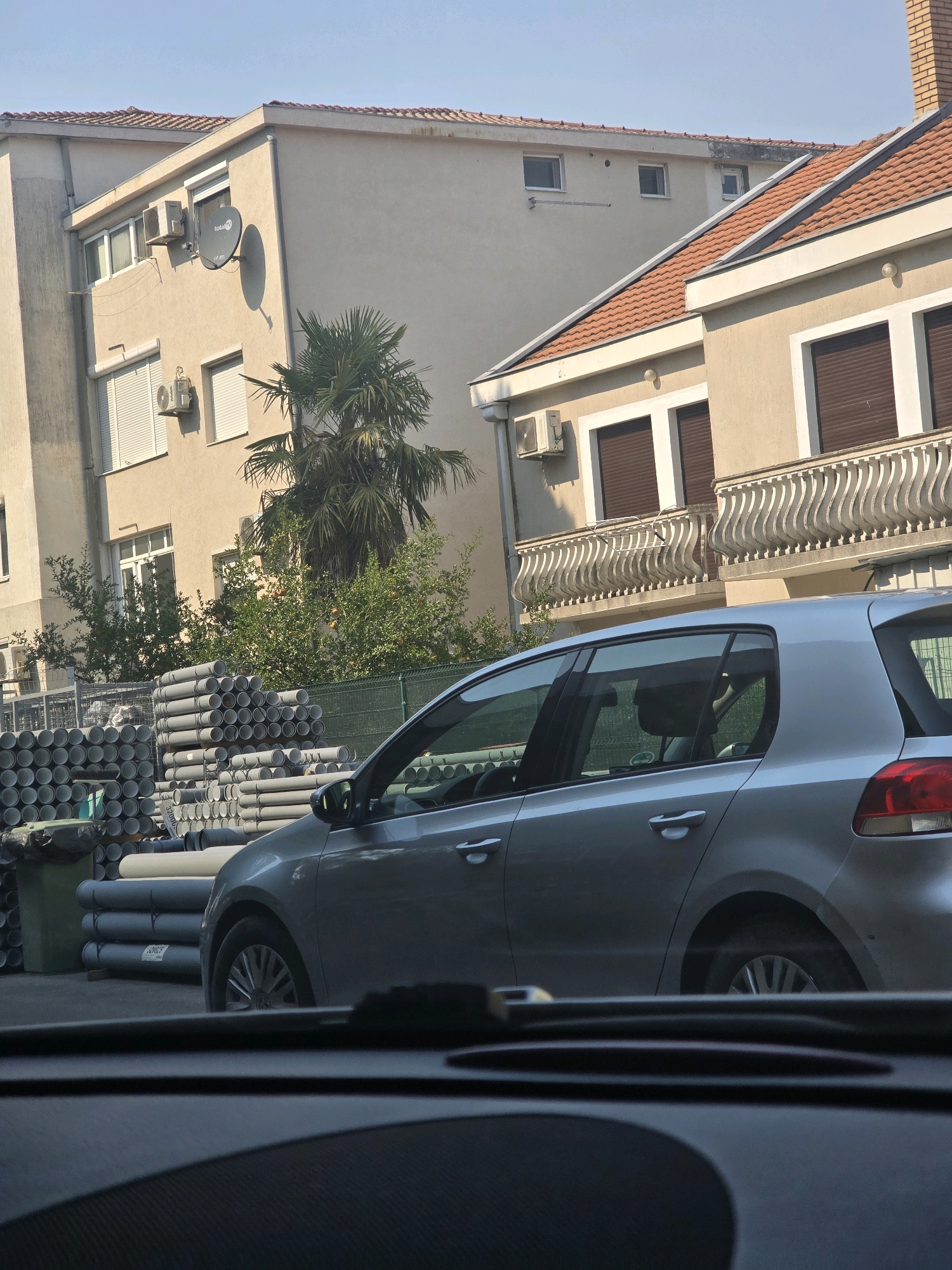 The image, taken in Podgorica, Montenegro, captures a street scene featuring two apartment buildings and a silver car. The building on the left is a light beige, multi-story structure with several windows, air conditioning units, and a satellite dish. The building on the right, with a red-tiled roof, also has air conditioning units and balconies with ornate railings. In front of the buildings, stacks of grey plastic pipes are visible, suggesting a construction or supply area. A palm tree and other greenery are interspersed between the buildings and the pipes. The silver car, a hatchback, is parked in front of the pipes, partially obscuring the view of the buildings behind it. The overall impression is a typical urban environment in the city, with a mix of residential architecture and commercial activity.