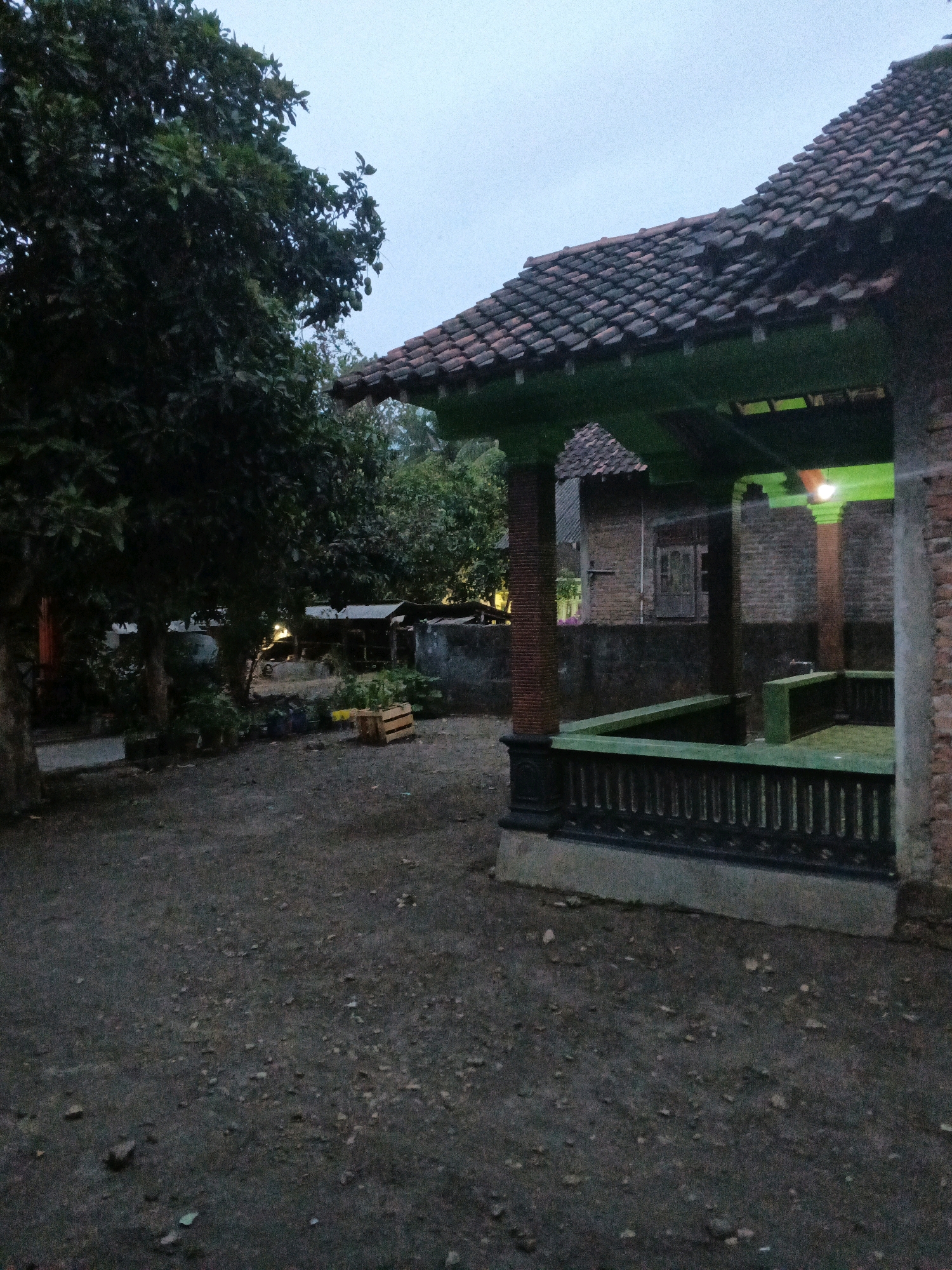 The image depicts an outdoor scene at dusk or twilight in Wates, Indonesia. In the foreground, a dirt and gravel yard occupies the lower portion of the frame. To the right, a raised, enclosed structure with a green railing and a tiled roof is visible. This structure appears to be a small pavilion or a memorial site, with brick pillars supporting a green-painted overhang. A light fixture emits a greenish glow from within the pavilion. Behind the pavilion, a brick building with a window is partially visible. To the left, a large tree with dark foliage dominates the scene. Further in the background, other trees and a low-lying structure with a corrugated roof are present. The overall atmosphere is somber and quiet, characteristic of a rural or suburban Indonesian setting in the evening. There are no people or animals visible in the image, and no specific actions are taking place.
