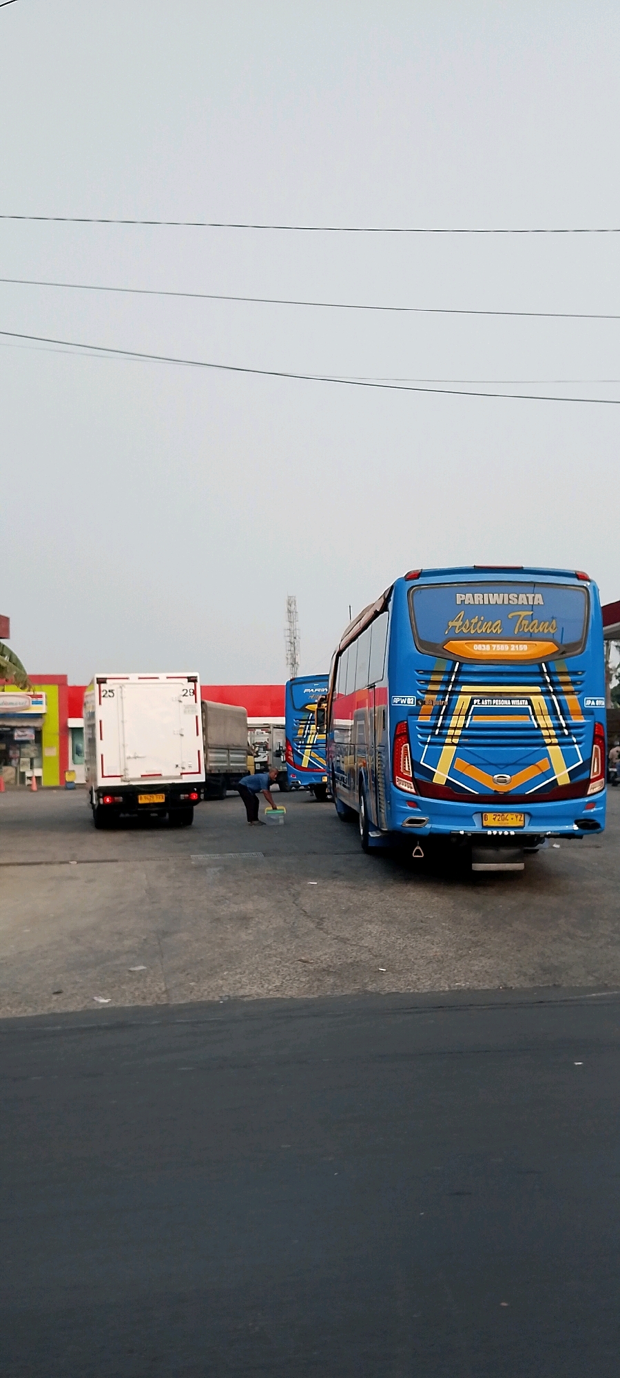 The image depicts an outdoor scene in the city of Bekasi, Indonesia. In the foreground, the asphalt surface of a parking lot or road is visible. In the mid-ground, there are several vehicles, including a white box truck with the numbers "25" and "29" on its back doors, and two blue passenger buses, one in front of the other. The bus in the foreground has "PARIWISATA Astina Trans" written on its rear, along with a phone number. The license plate of this bus reads "B 7204 YZ". A person in a blue shirt and dark pants is bending down, holding a clear plastic container. In the background, there are buildings with red and yellow accents, possibly a gas station or convenience store. Overhead, power lines stretch across the frame against a pale sky. The overall environment appears to be a commercial or transportation hub.