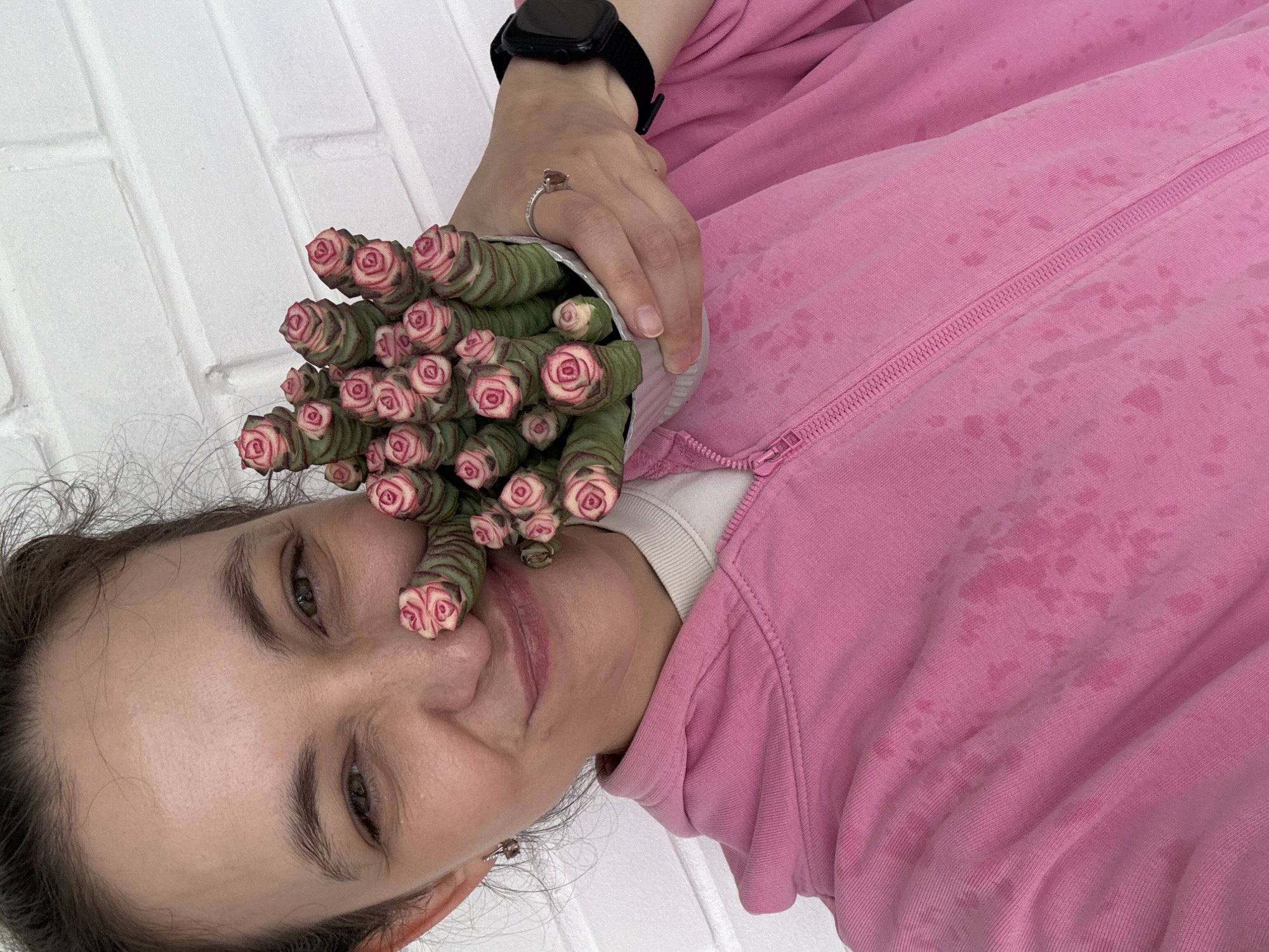 In an outdoor urban setting, against a white brick wall, a woman with brown hair pulled back and green eyes poses for a close-up selfie. She is wearing a pink hooded sweatshirt with a zipper. In her right hand, she holds a small potted succulent plant, which is positioned near her face, partially obscuring her nose. The succulent's leaves are stacked in a spiral pattern, resembling small pink roses. Her left hand rests on her chest, showcasing a silver ring on her ring finger and a black smartwatch on her wrist. The scene is brightly lit, suggesting daylight. There are no discernible landmarks that would place this image in Chaikovsky, Russia; it could be any location with a brick wall.