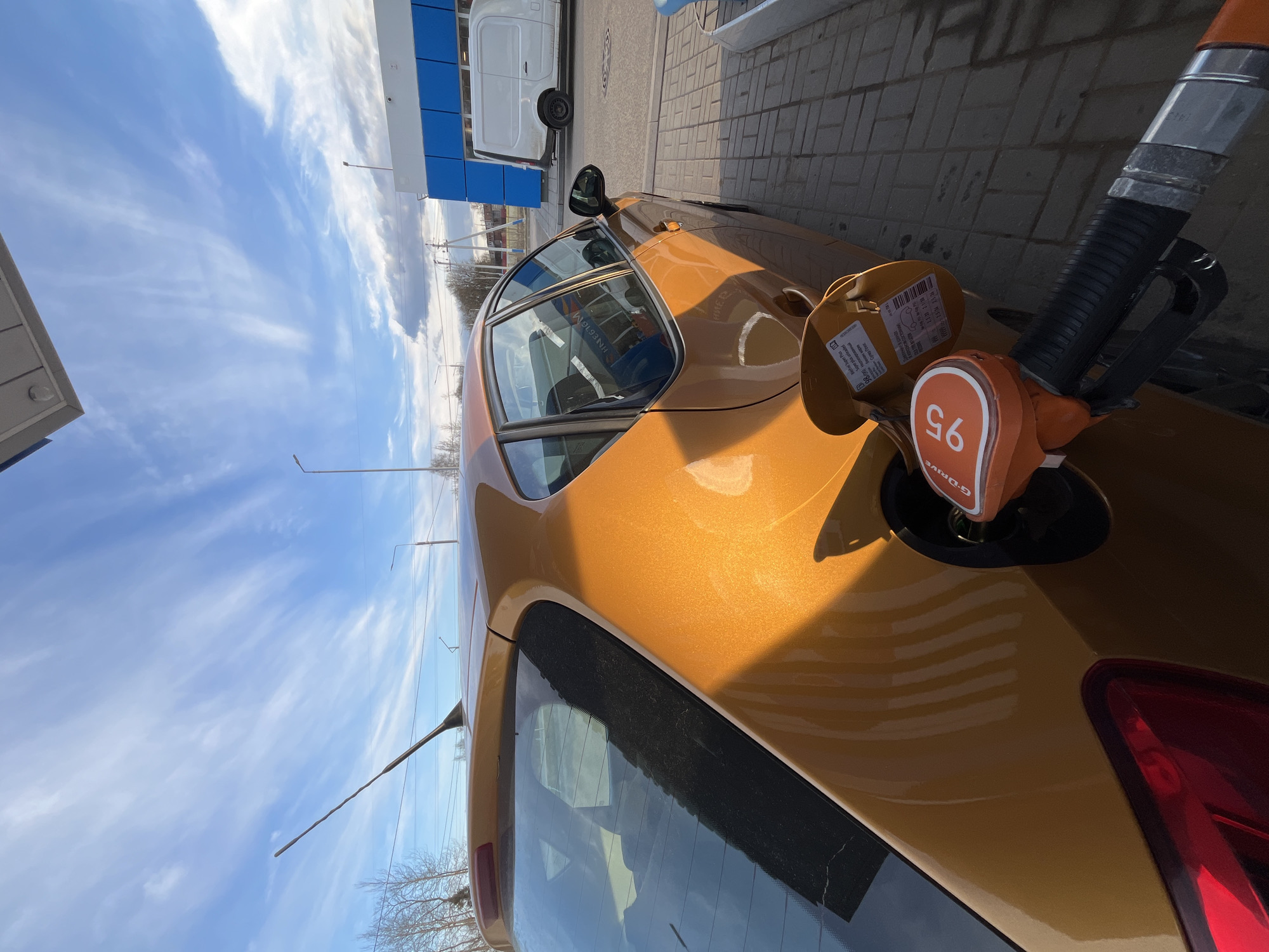 Here is a detailed description of the image:
**Scene**: An eye-level, outdoor shot depicts a golden-colored car being fueled at a gas station.
**Subjects/Objects**:
- A gas pump nozzle, marked with "95" and the word "G-DRIVE" written over it, is inserted into the car's gas tank. Text on the gas tank cover mentions “Super Plus unleaded” in both Russian and English.
- The car, in a metallic golden hue, dominates the view.
- In the background, a white van is parked near a building with blue and white paneling, likely part of the gas station structure.
- The sky is visible, with some wispy clouds, suggesting fair weather.
**Environment**: The setting appears to be a gas station forecourt, with paved ground visible around the car and gas station structures.
**Location**: Based on the translated text on the gas tank and the car, the location is Klin, Russia.