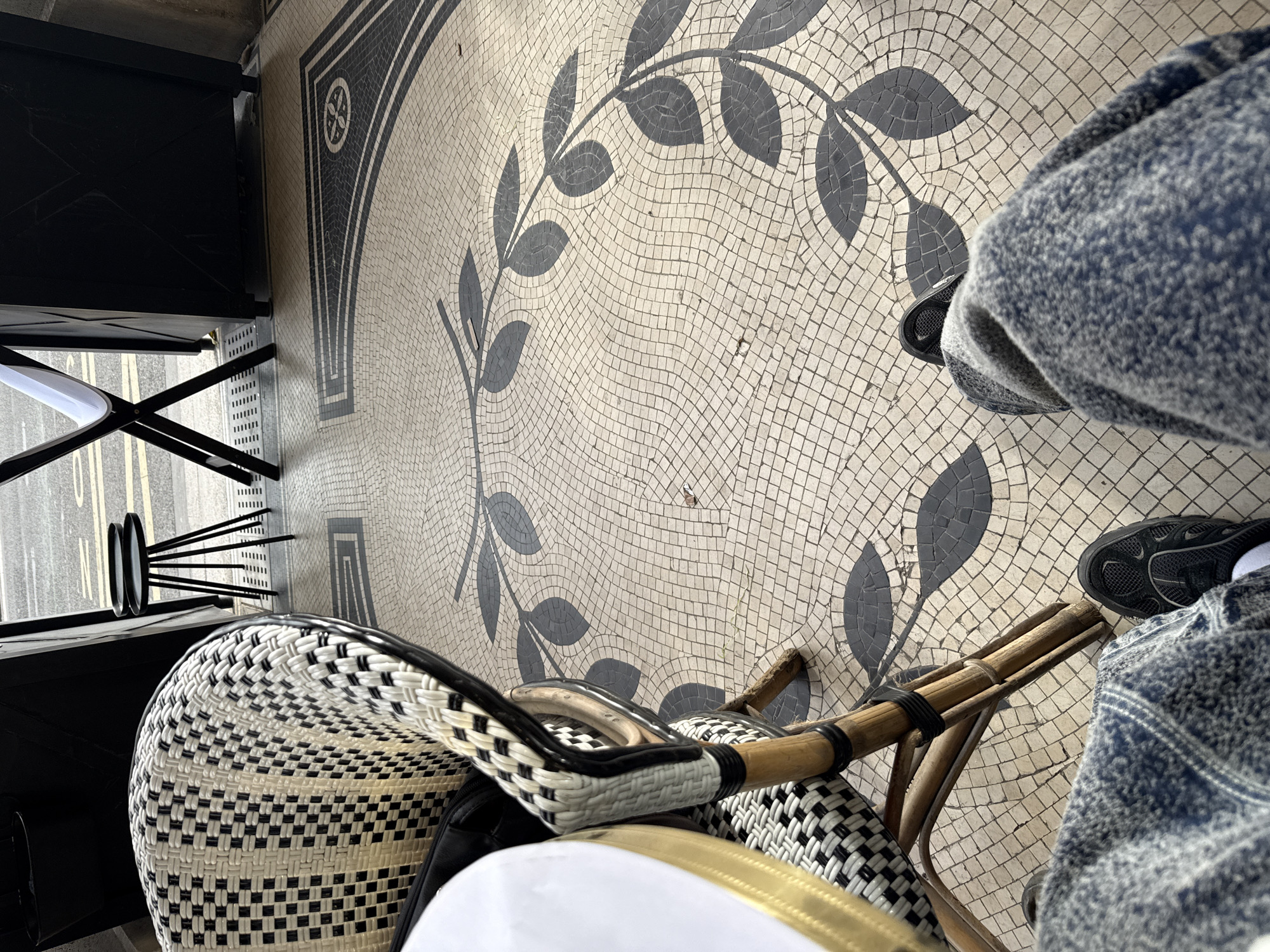 The image shows a close-up, high-angle view of a café scene in Paris, France. The ground is covered in a decorative mosaic tile pattern with a laurel wreath design. A table with black metal legs and a white top is positioned on the left side of the image. There is a chair with a woven black-and-white seat and a bamboo frame. The legs of a person wearing jeans and black sneakers are visible on the right side of the image, suggesting the person is seated. The focus is on the texture and patterns of the tiled floor and the woven chair, creating a sense of being in a Parisian café.