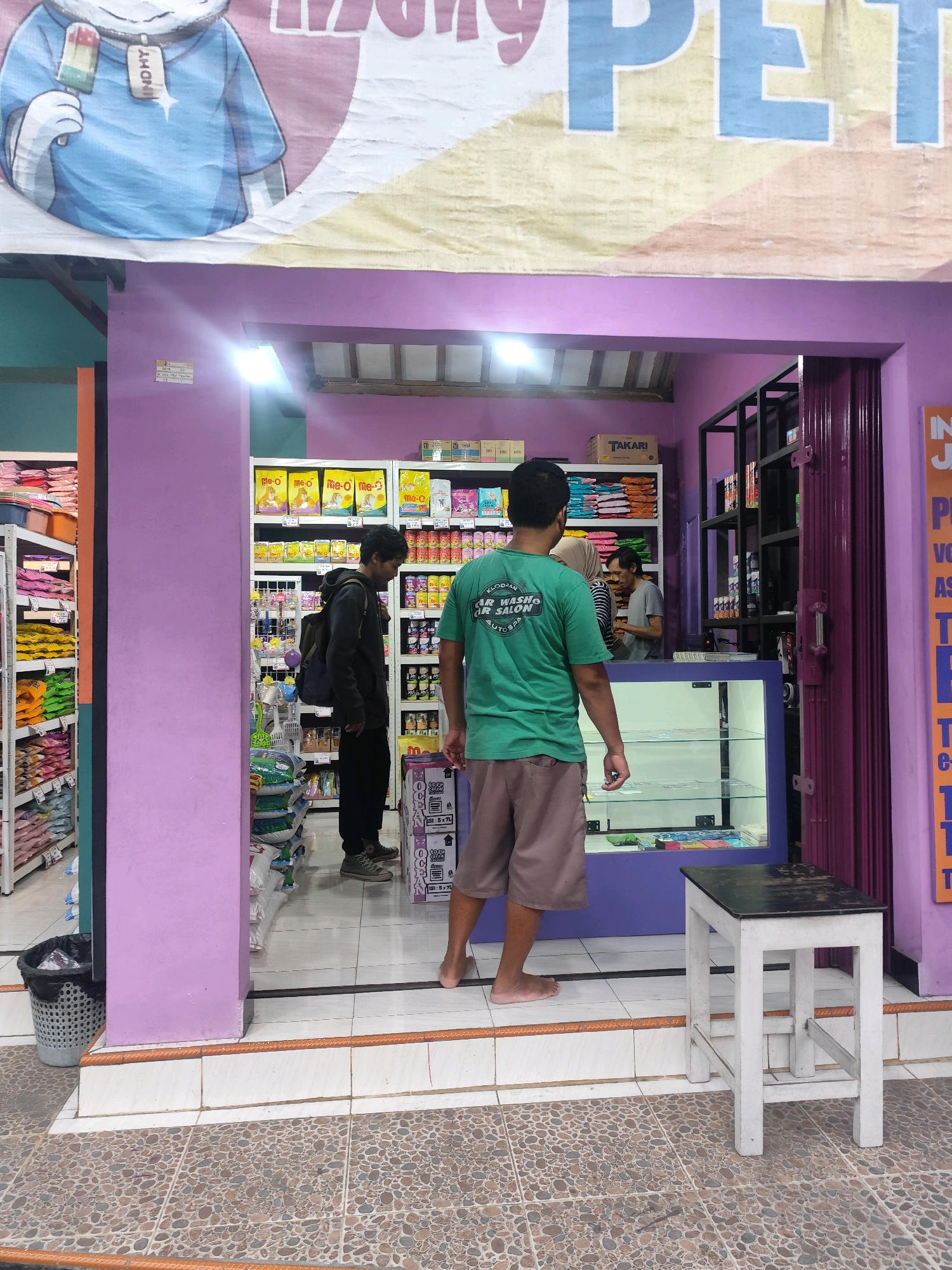 A medium shot captures the entrance of a small shop in Bambanglipuro, Indonesia. The shop, painted a vibrant purple, is open to a tiled courtyard. Inside, shelves are stocked with various goods, including what appear to be pet food bags and other packaged items. A young man wearing a dark hoodie and carrying a backpack stands near the shelves, facing them. In front of him, another man in a green t-shirt and brown shorts is facing away from the camera, looking towards the interior of the shop. A glass display case is visible in the shop's interior. To the right of the entrance, a narrow aisle leads deeper into the shop, where more shelves and a folded metallic door are present. Outside the shop, a small white stool with a black top sits on the tiled ground. A black trash bin is positioned near the left side of the shop entrance. The scene appears to be well-lit by artificial lights.