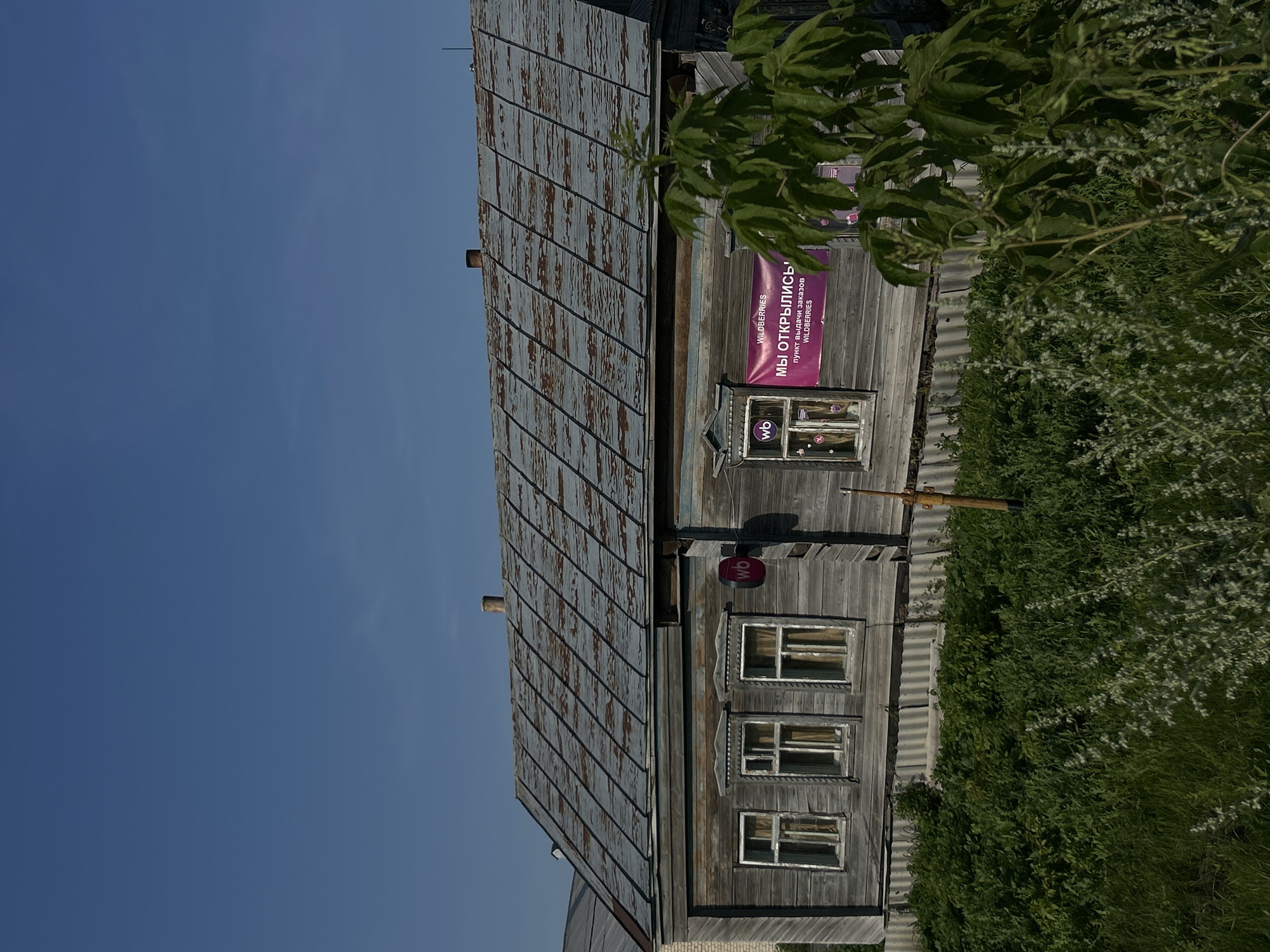 The image depicts a weathered, wooden, two-story building with a corrugated metal roof. The building is situated in an outdoor environment with a clear blue sky overhead and lush green vegetation in the foreground. Several windows are visible on the building's facade, some adorned with decorative trim. A pink banner with Russian text and the "wb" logo is prominently displayed on the right side of the building. There are no people visible in the image. The overall impression is one of rural or small-town architecture, possibly in a setting such as the city of Mazan, Russia, given the context.