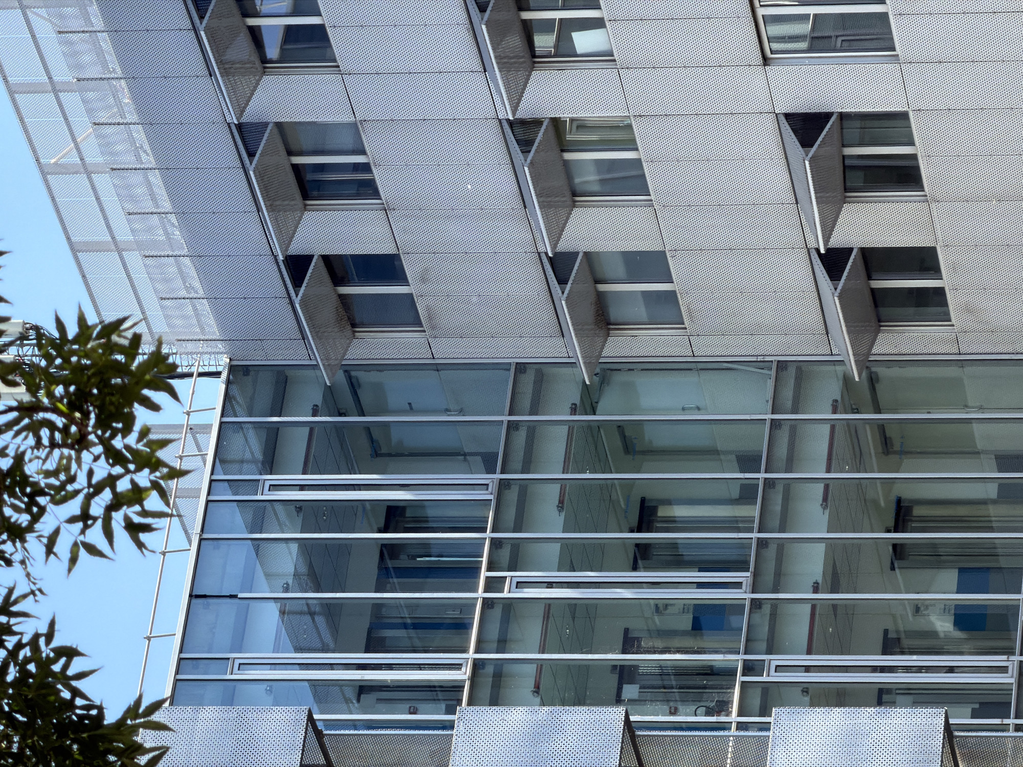 The image shows the exterior of a modern building with a glass facade on the left and a metal facade with adjustable sunshades on the right. The building appears to be in an urban setting, as indicated by the blue sky visible above and the presence of trees in the foreground. The glass facade is composed of large vertical panels framed by metal mullions, revealing the interior of the building, which includes elevators and office spaces. The metal facade is made of perforated panels and features several windows, each equipped with angled sunshades that are partially open. The overall impression is one of contemporary architecture, characterized by clean lines and industrial materials. This building is located at the Institut Lumière in Lyon, France.