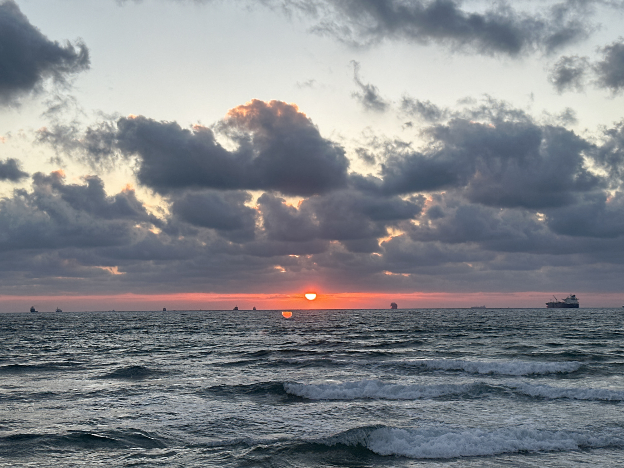 The image depicts a serene sunset over the Mediterranean Sea, likely near Haifa, Israel. The sky is filled with dramatic, dark clouds tinged with warm orange and pink hues from the setting sun, which is partially visible just above the horizon. Below the vibrant sky, a calm sea stretches out, with gentle waves rolling towards the shore. Several cargo ships are scattered across the water, silhouetted against the colorful sky, suggesting an active port or shipping lane. The overall mood is one of tranquility and awe, capturing the natural beauty of a coastal sunset. The time of day is clearly evening, as indicated by the sunset and the soft light. The weather appears to be clear enough for shipping, though the clouds add a dramatic element. There is no visible text in the image.