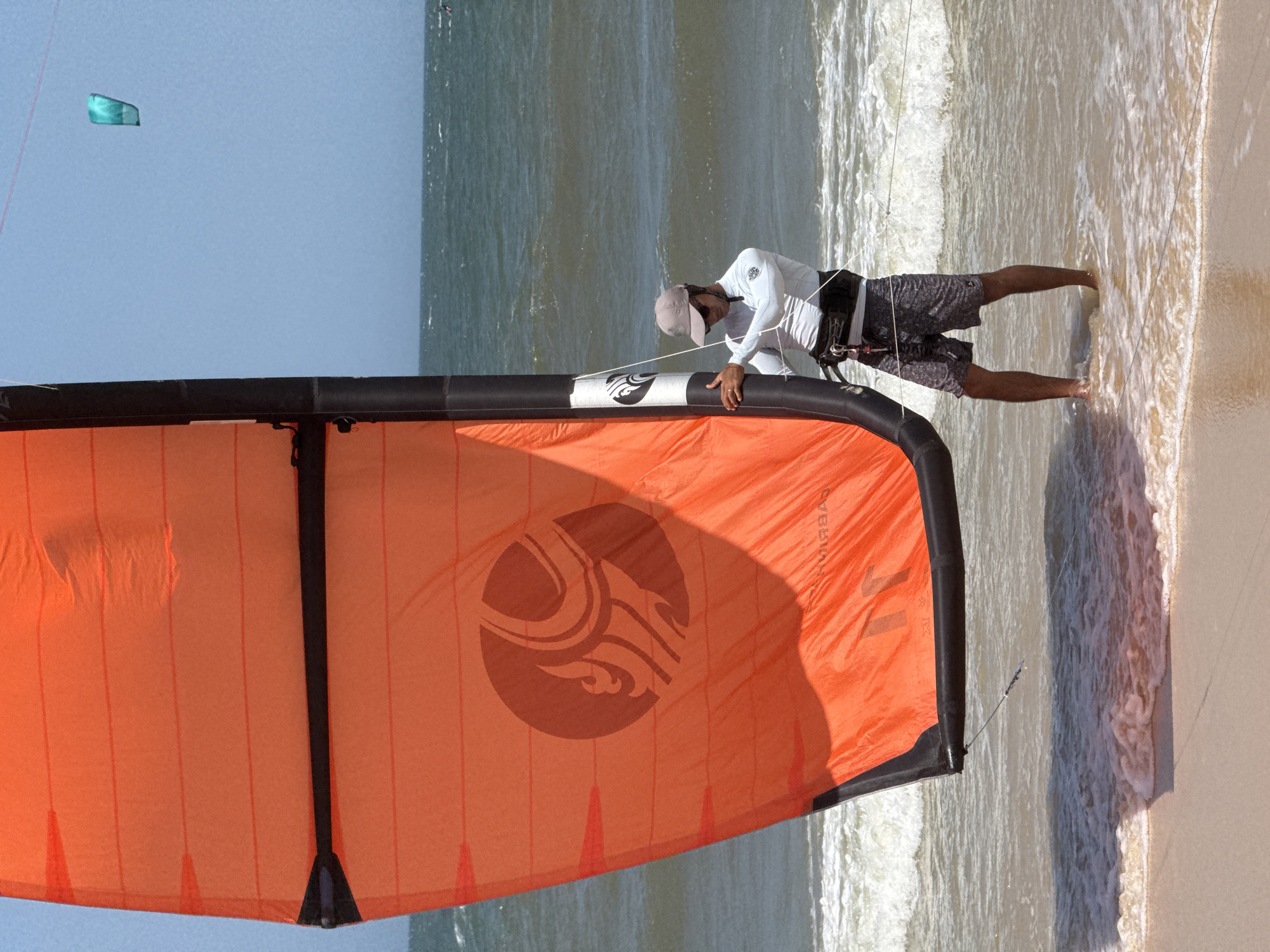 A person stands in shallow ocean water on a sandy beach, holding a large orange kite. The kite is partially unfurled and has a black leading edge with white accents. The person is wearing a light-colored long-sleeved shirt, dark patterned shorts, a grey baseball cap, and sunglasses. They are also wearing a kiteboarding harness around their waist. The water is choppy, with waves breaking on the shore. In the distance, the ocean stretches to the horizon under a clear blue sky. Another smaller, teal-colored kite is visible high in the sky to the right of the main kite. This scene is located in Mũi Né, Vietnam, a popular destination for kitesurfing.
