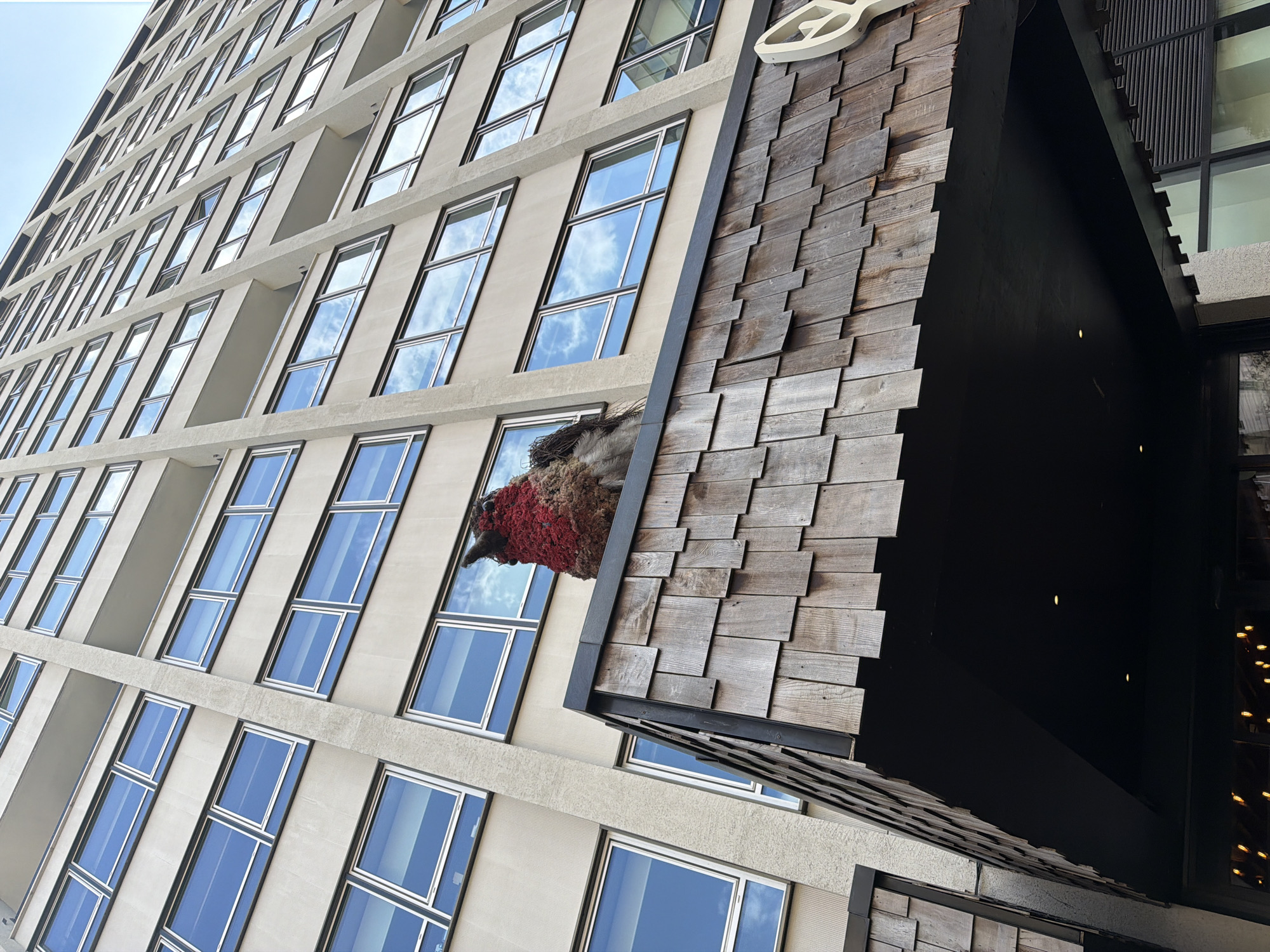 The image presents a striking upward perspective of a modern architectural ensemble in an urban setting, identified as London, United Kingdom. The left portion of the frame features a tall, contemporary building with a light-colored, possibly concrete or stone, facade. This building is characterized by numerous uniformly sized rectangular windows, arranged in a grid-like pattern across multiple stories. These windows brightly reflect a clear or partly cloudy blue sky, with distinct white cloud formations visible in the glass.
Integrated with and adjacent to this main structure, on the right side of the image, is a unique, dark-colored architectural feature. This element has a steeply sloped surface covered in a mosaic of weathered wooden shingles, creating a rustic and textured contrast to the smooth modern facade. From an opening within this shingled structure, at about mid-height, protrudes a large, sculptural bird's head and neck. The bird's head is distinctly reddish-brown, appearing to be crafted from natural, possibly organic materials like moss or fibers, while its neck section displays a more earthy brown texture. This prominent art installation extends outwards, facing the windows of the main building.
The scene is illuminated by bright daylight, indicated by the clear sky reflections and well-lit surfaces. No people or visible text are present. The image captures an intriguing blend of modern architecture and naturalistic art, forming a notable feature within the city environment.
