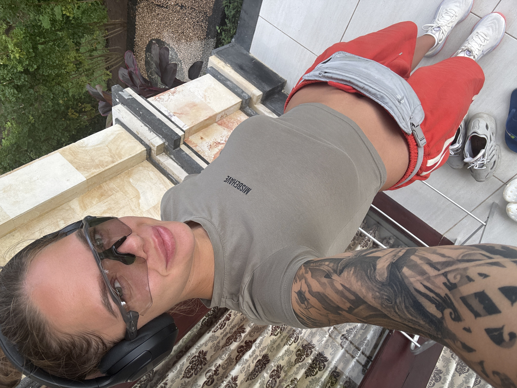 A selfie taken from a high angle depicts a person in Unknown, Indonesia. The individual, identifiable as female, is clad in a gray short-sleeved top with "MISBEHAVE" printed on it, red shorts with white stripes, and white sneakers with red accents. A gray fanny pack is slung across their midsection. They are also wearing black headphones and sunglasses. Their left arm is heavily tattooed. The backdrop includes a patterned surface, possibly a curtain or table, and a white-tiled floor area. Beyond the floor, there's a view of trees and other greenery suggesting an outdoor environment. The architecture in the background includes a white-tiled wall or railing. The image is captured at eye-level, suggesting the individual is holding the camera or phone at arm's length.