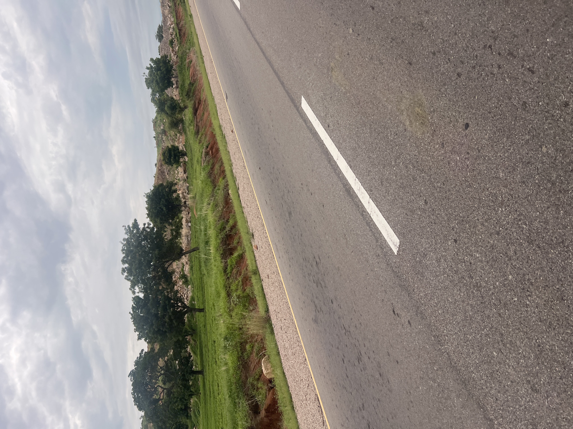 The image depicts a roadside scene on an overcast day. A paved road with a white dividing line and a yellow edge line runs diagonally across the frame from the lower left to the upper right. To the left of the road, a grassy embankment slopes upwards, with patches of red soil visible. Several trees with green foliage are spaced along the top of the embankment, and beyond them, the terrain appears rocky and sparsely vegetated. The sky is filled with grey clouds. The scene is devoid of people or any visible activity, suggesting a quiet, rural or semi-rural setting, consistent with an area like Minda, Nigeria. The lighting conditions indicate it is daytime, likely mid-morning or afternoon.