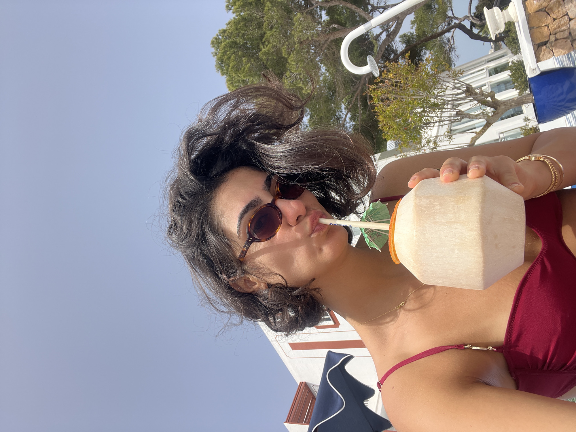 A young woman wearing a maroon-colored bikini and brown sunglasses is seen drinking from a white coconut with a green straw. She is wearing gold bracelets on her left wrist and a gold necklace. Her hair is short and wavy. The background includes a tree, part of a white building with red trim, a white shower fixture, and a partial view of a blue and white umbrella. The location is Santa Eulària des Riu, Spain.