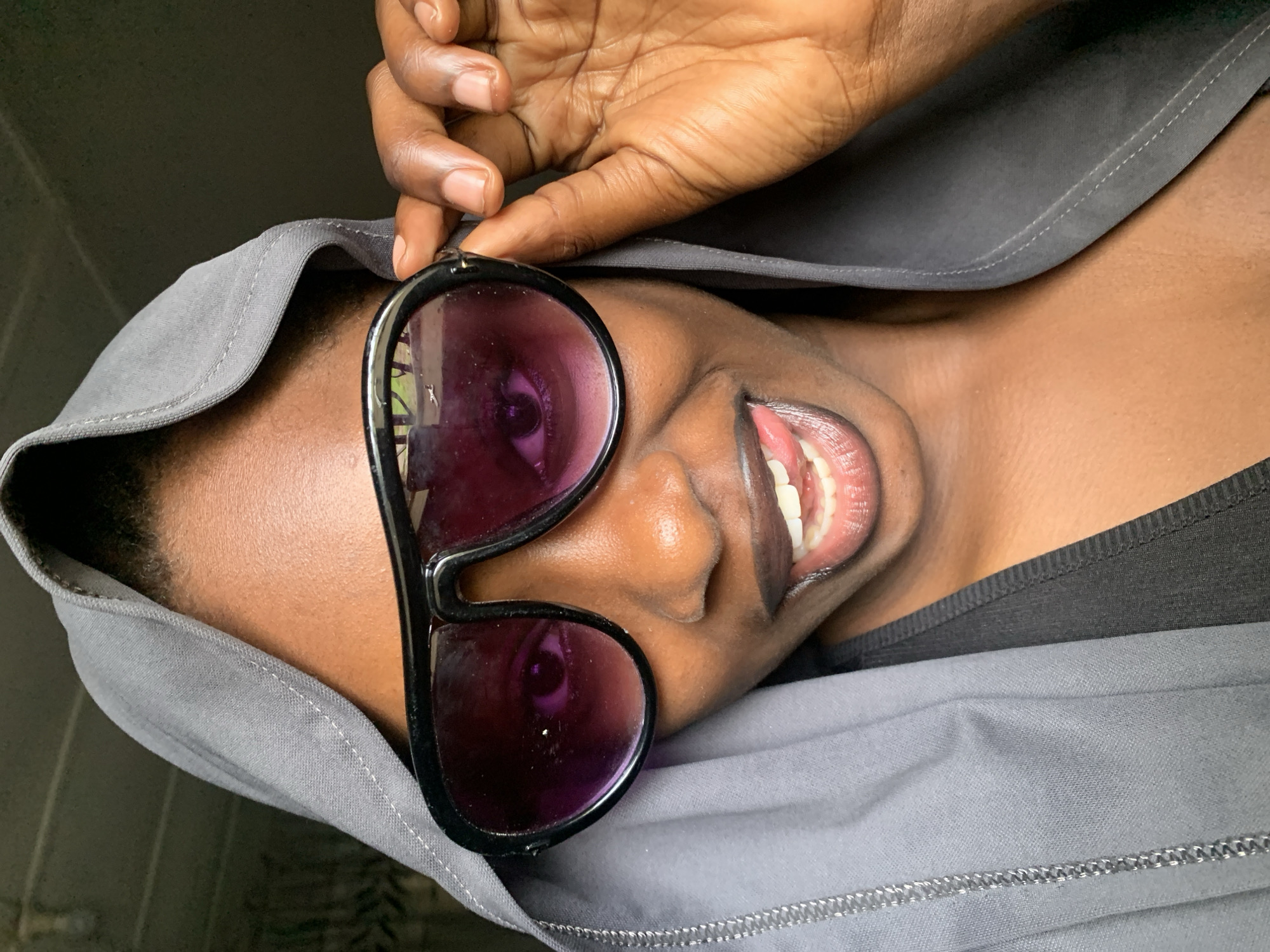 The image is a close-up, vertical, portrait-style photograph of a person wearing sunglasses and a grey hooded garment. The subject's face is turned to the side, with their eyes visible through the purple-tinted lenses of the sunglasses. Their mouth is open, revealing teeth and tongue, with a playful or joyful expression. A hand with dark skin is seen adjusting the sunglasses, with fingers delicately positioned on the frame. The grey fabric of the hood is prominent, framing the subject's face and obscuring parts of their head and shoulders. The lighting appears to be natural, suggesting an indoor or outdoor setting during the daytime. There is no visible text in the image. The context provided, "Rafin Par, Nigeria," suggests the location of the photograph.