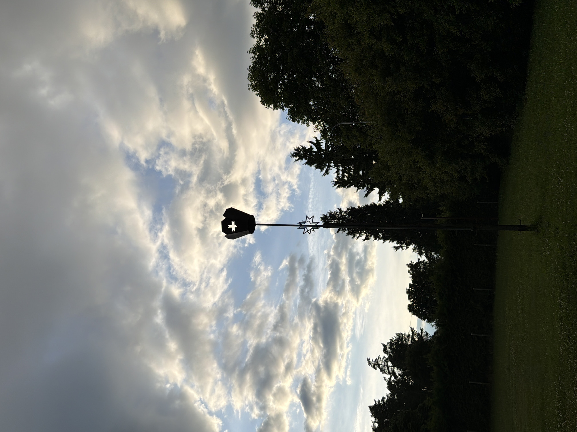 A tall, slender lamppost with a decorative, star-shaped cutout at the top stands in a grassy field. The lamppost is silhouetted against a cloudy sky. In the background, a dense line of trees and bushes forms a dark border. The ground is covered in green grass, with a few scattered white wildflowers visible. The scene is lit by the soft glow of what appears to be late afternoon or early evening light filtering through the clouds. The overall impression is one of a peaceful, somewhat overcast outdoor setting in the city of Ulbroka, Latvia.